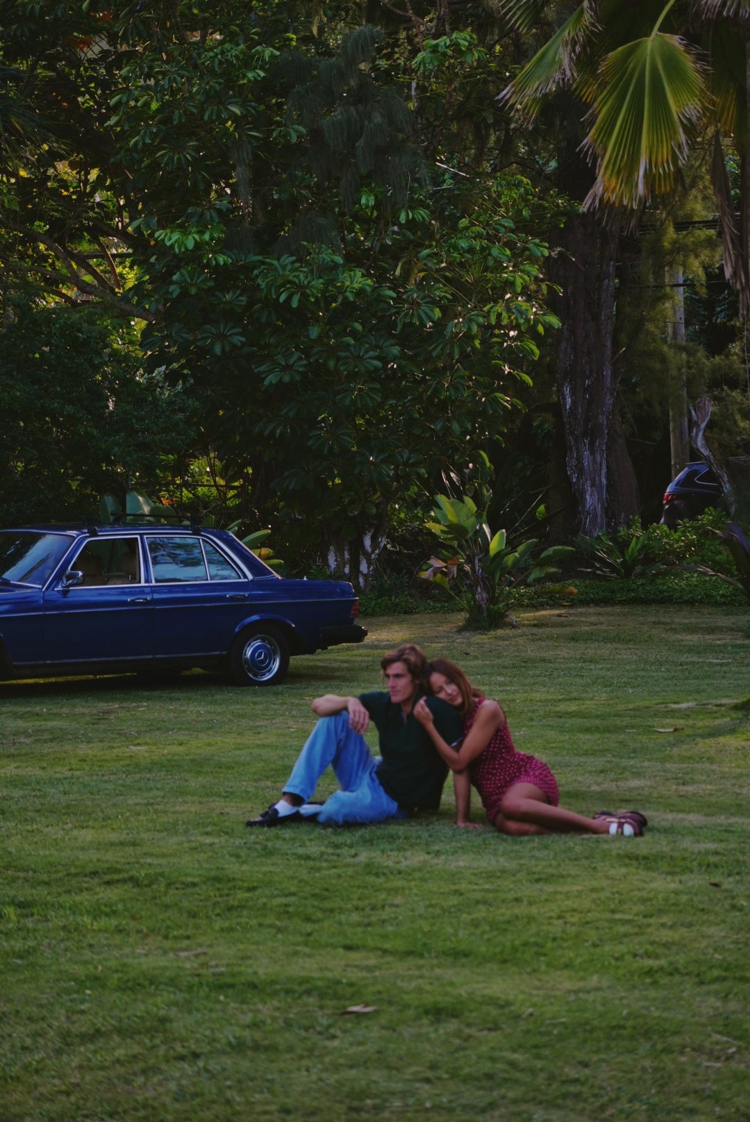 A man and a woman sitting on grass in a park with trees and vintage cars in the background. The woman leans her head on the man's shoulder. The setting appears to be late afternoon or early evening.