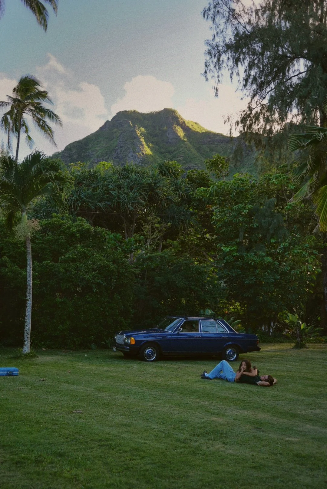 A tropical landscape with lush green trees and a mountain in the background. Two women are lying on the grass near a vintage blue car, enjoying the scenery.