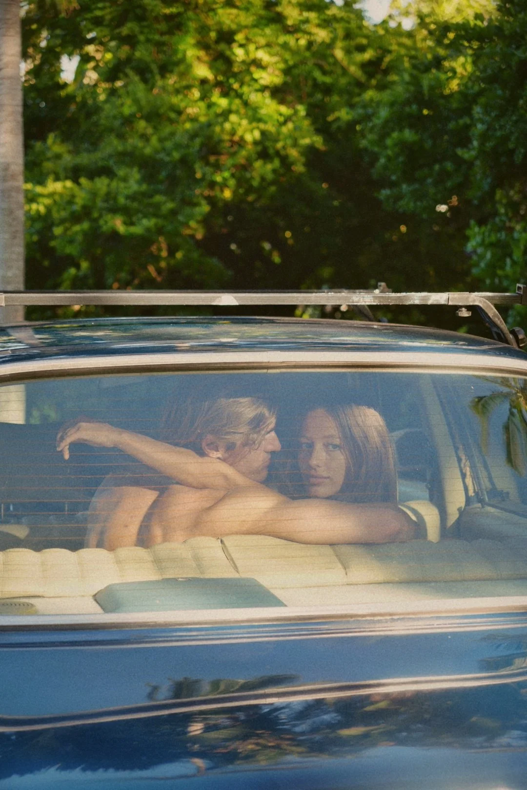 A couple, a man and a woman, sitting inside a car looking at each other through the rear window, with trees and foliage in the background.