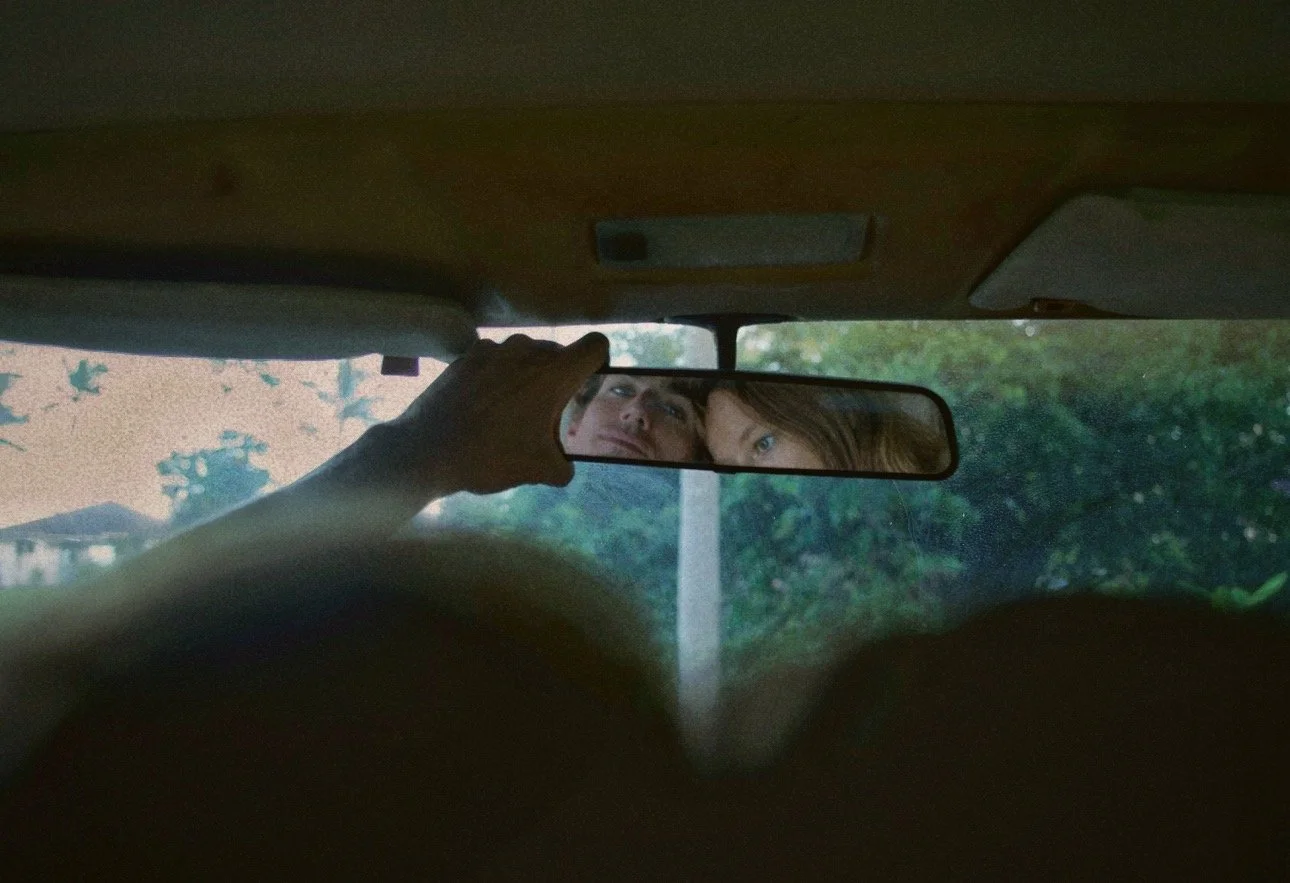 A person adjusting the rearview mirror inside a vehicle, with two people visible in the mirror's reflection, outdoors with trees and a building in the background.