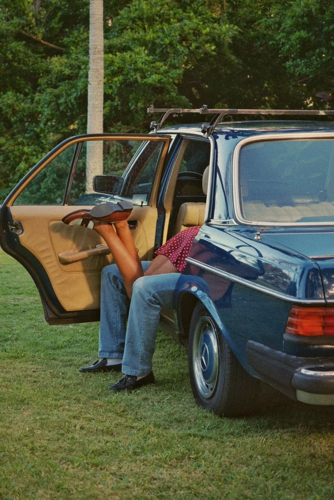 A person with their legs stretched out, lying across the front seats of a parked blue car with its driver's door open, on a grassy area with green trees in the background.