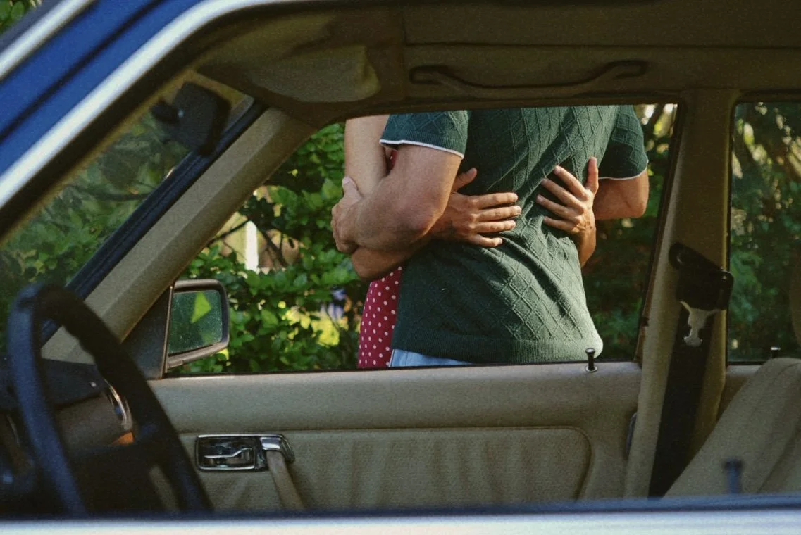 A person hugging another person outside a car, seen through the car window, with greenery in the background.