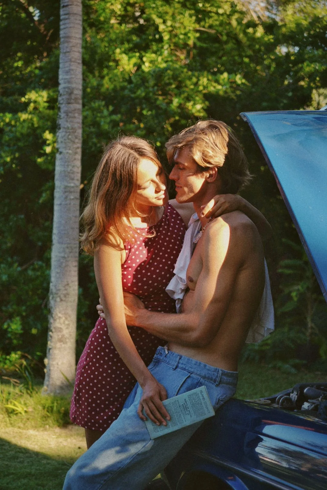 A man and woman embrace outdoors near a blue car on a sunny day, surrounded by trees.