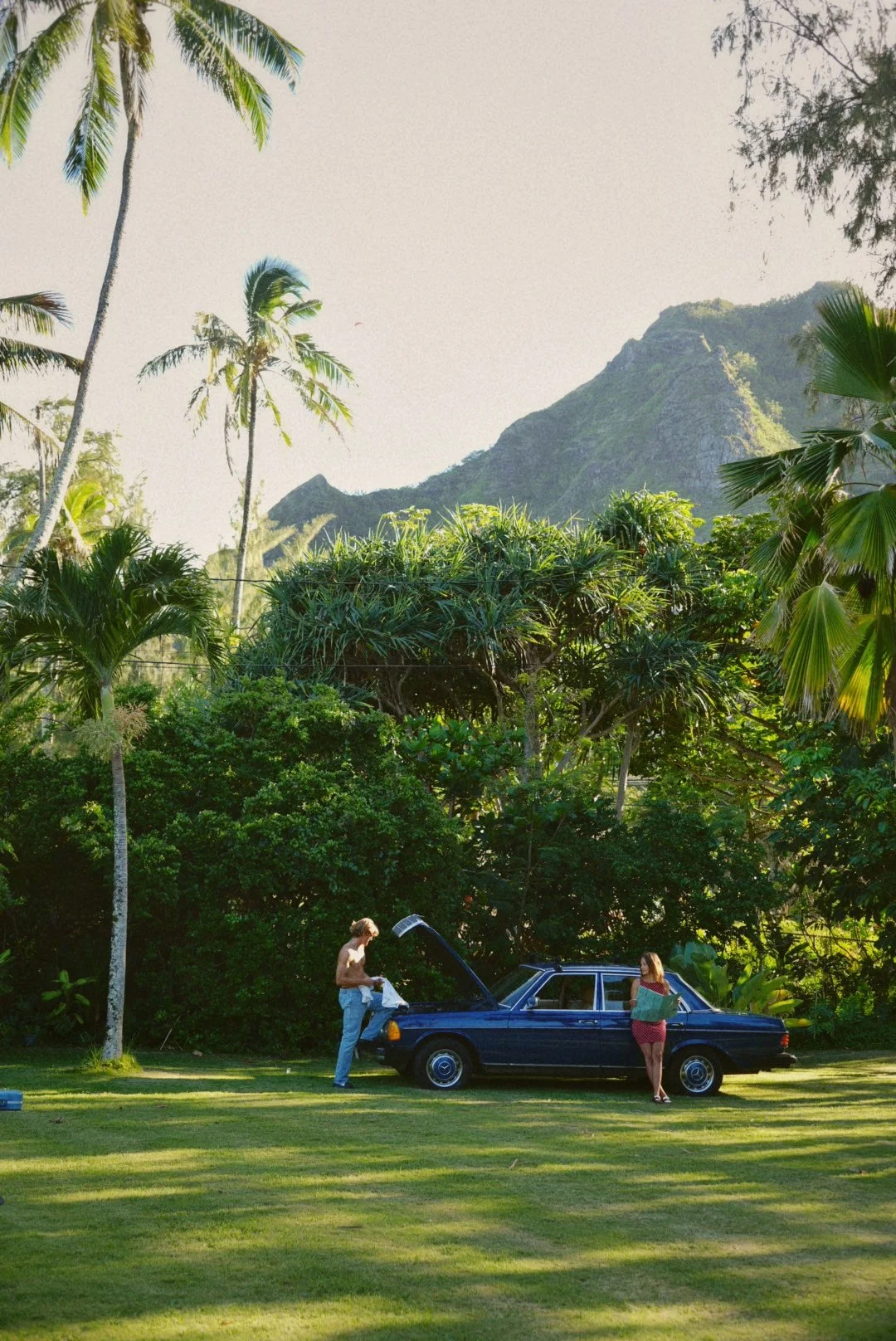 A man and woman outdoors near a blue car with its hood open, surrounded by lush tropical trees and plants, with mountains and a clear sky in the background. The woman is looking at a map, and the man is holding a piece of cloth or clothing.