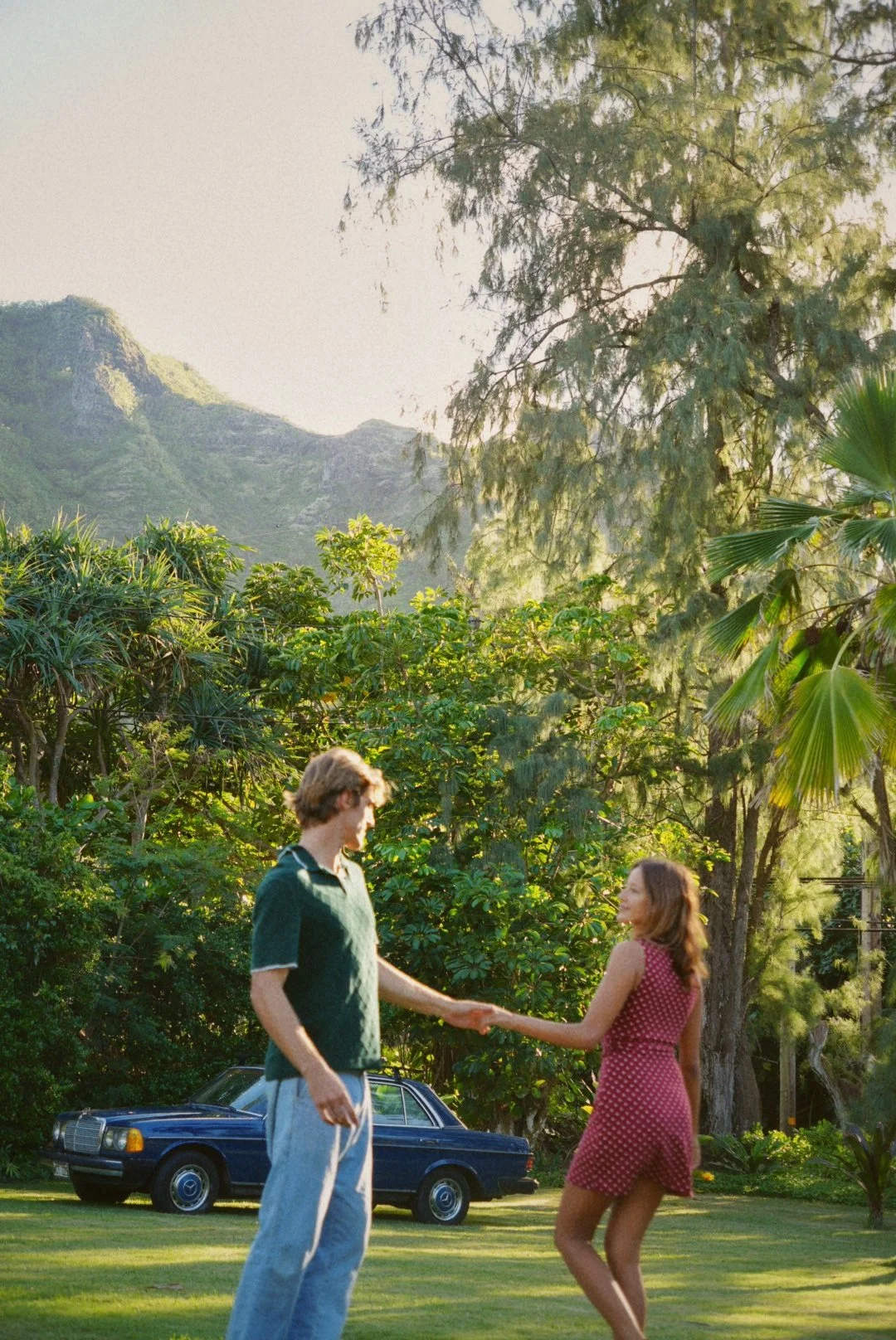 A young man and woman are holding hands and dancing in a lush green park with trees and mountains in the background. There is a vintage blue car parked on the grass nearby, and the scene is illuminated by warm sunlight.
