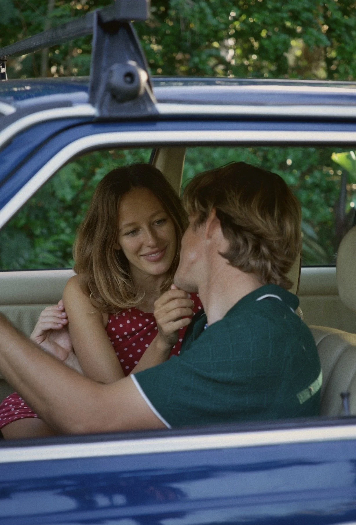A young woman and man sitting in a car, engaged in a romantic moment, with the woman smiling gently as the man touches her chin, against a background of green foliage.