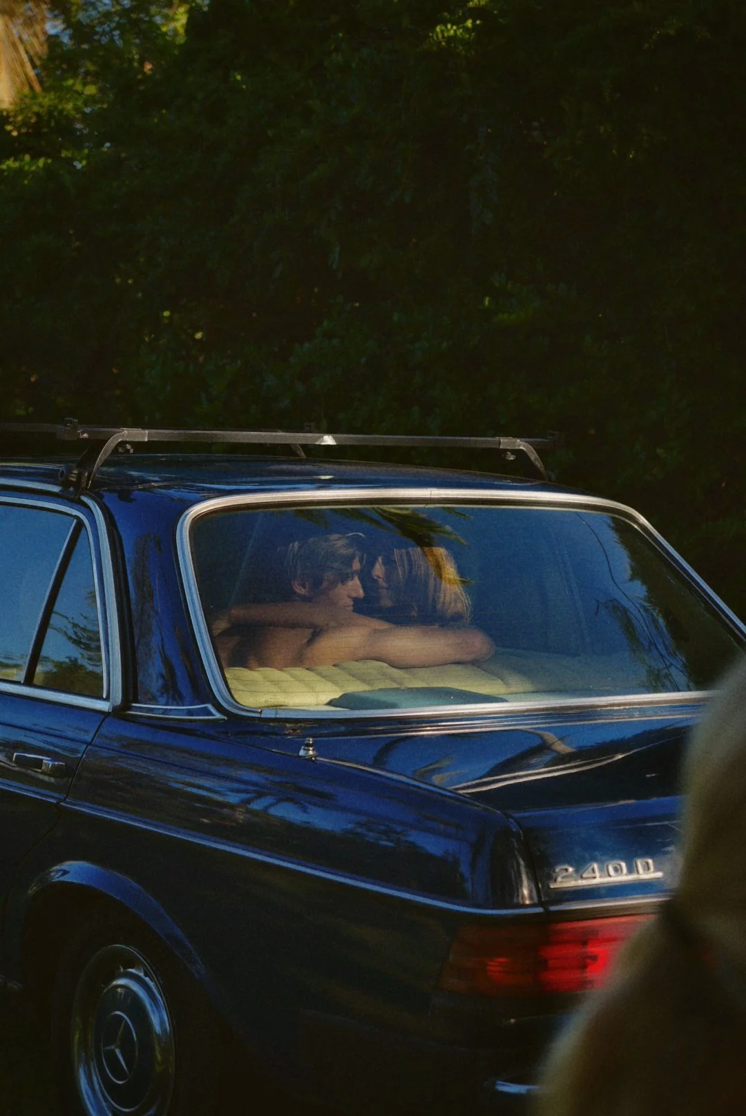 A blue vintage Mercedes-Benz 240 D car with two people inside, embracing each other, viewed through the windshield.