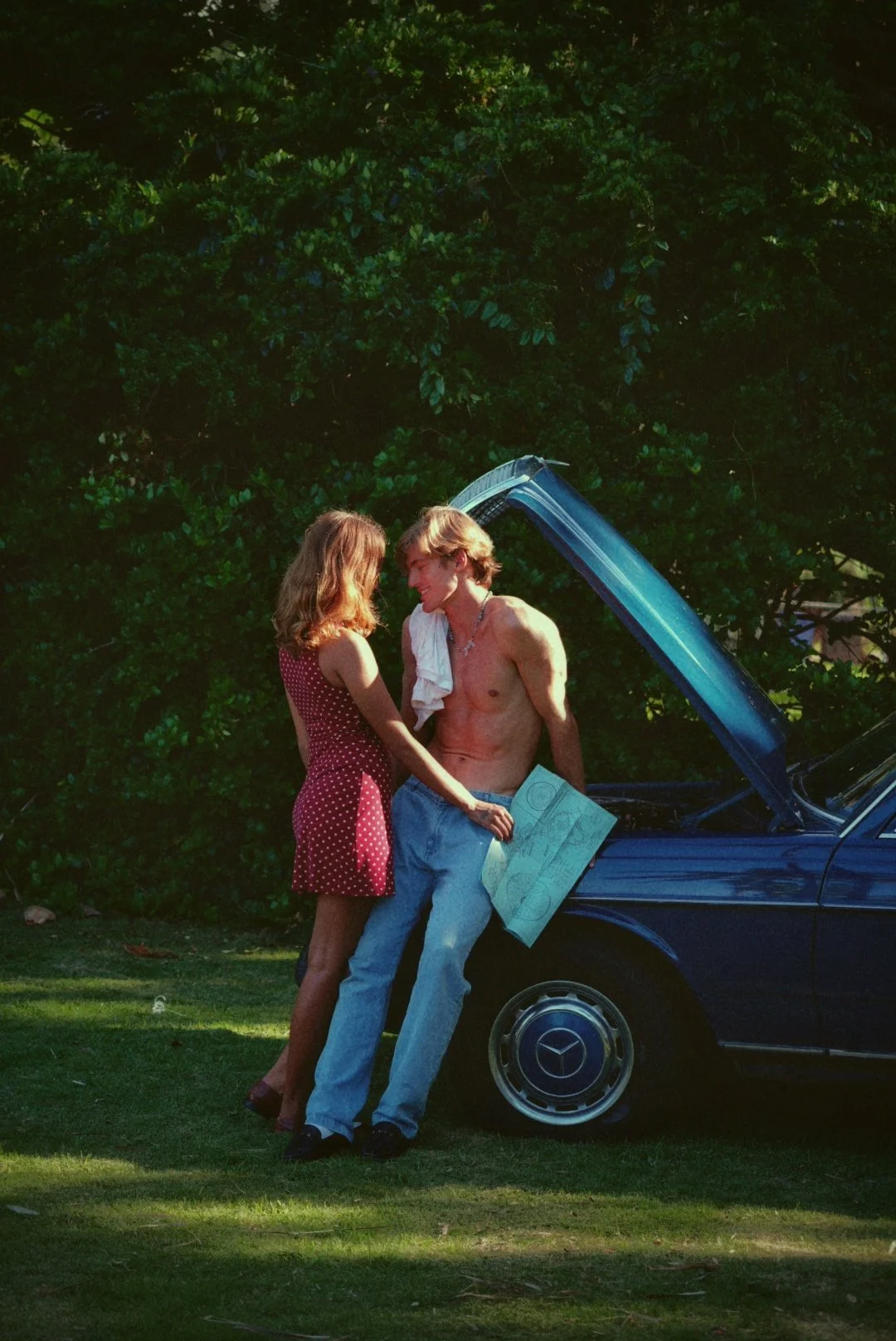 A shirtless man with a towel around his neck holding a blueprint leaning against a blue car with an open hood, a woman in a red polka dot dress standing close to him in a park-like area with green trees in the background.