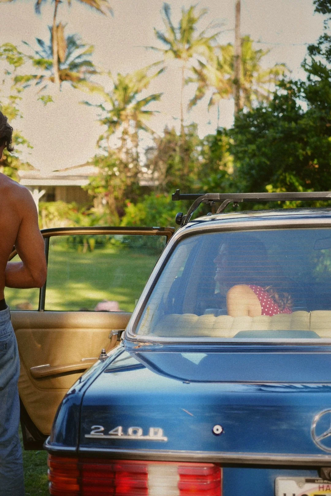 A blue vintage car parked outdoors with a woman sitting in the backseat and a shirtless man standing nearby with the car door open, surrounded by greenery and palm trees.