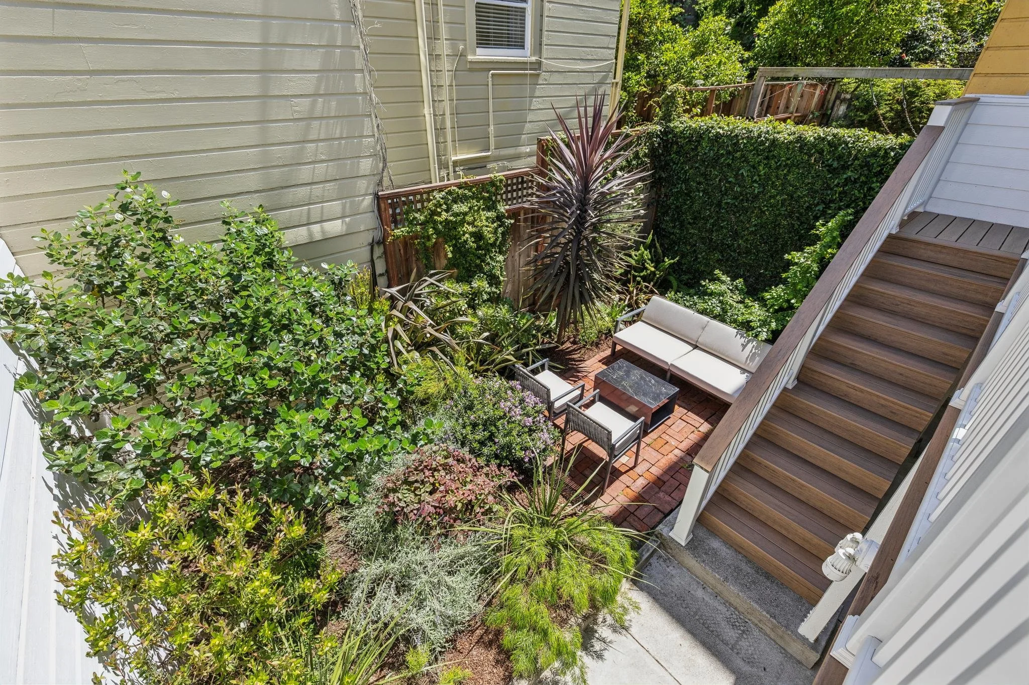 A small outdoor patio area with a white cushioned chair, a black table, and various green plants and shrubs. A staircase leads up to the house, and a wooden fence with greenery separates the yard from neighboring areas.