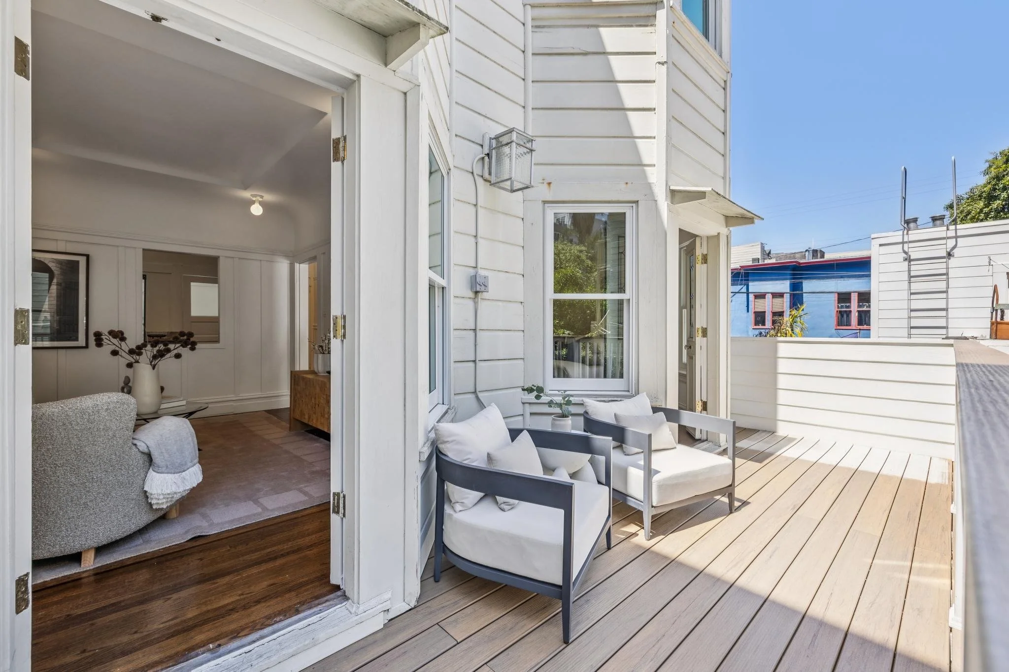 A cozy outdoor balcony with a white sofa and a gray armchair, decorated with white cushions, and a small wooden table with potted plants, overlooking a sunny sky and neighboring colorful buildings.