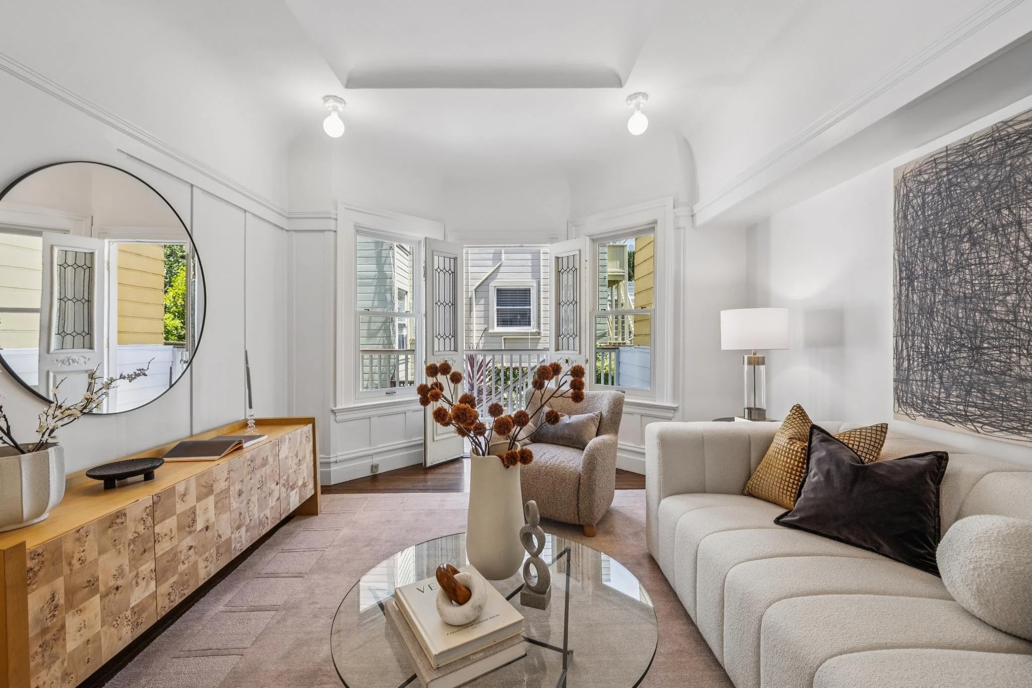 Living room with white walls, large windows, a beige armchair, a white sofa with gold and black pillows, a glass coffee table with decorative items, a framed abstract artwork, and a tall lamp.