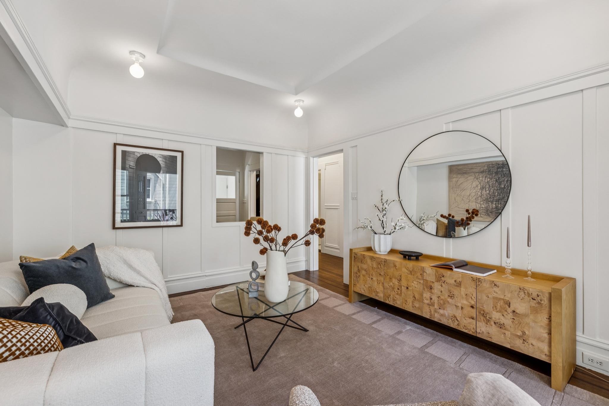 Living room with white walls, a white sofa, decorative pillows, a glass coffee table, a wooden sideboard with decorative items, a large round mirror, framed artwork, and a vase with dried flowers.