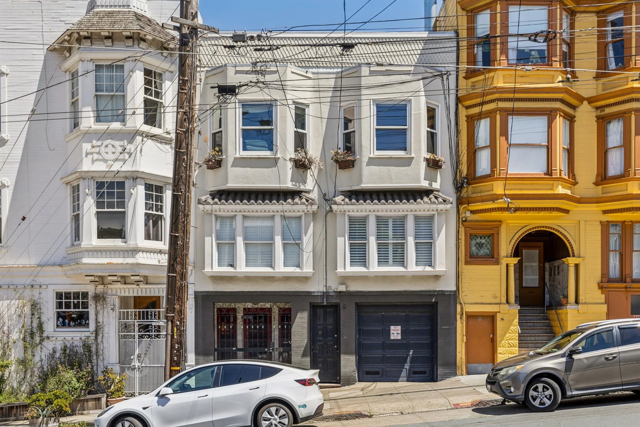 A row of colorful Victorian houses with bay windows and ornate details, parked cars in front, and a utility pole with wires overhead in an urban neighborhood.