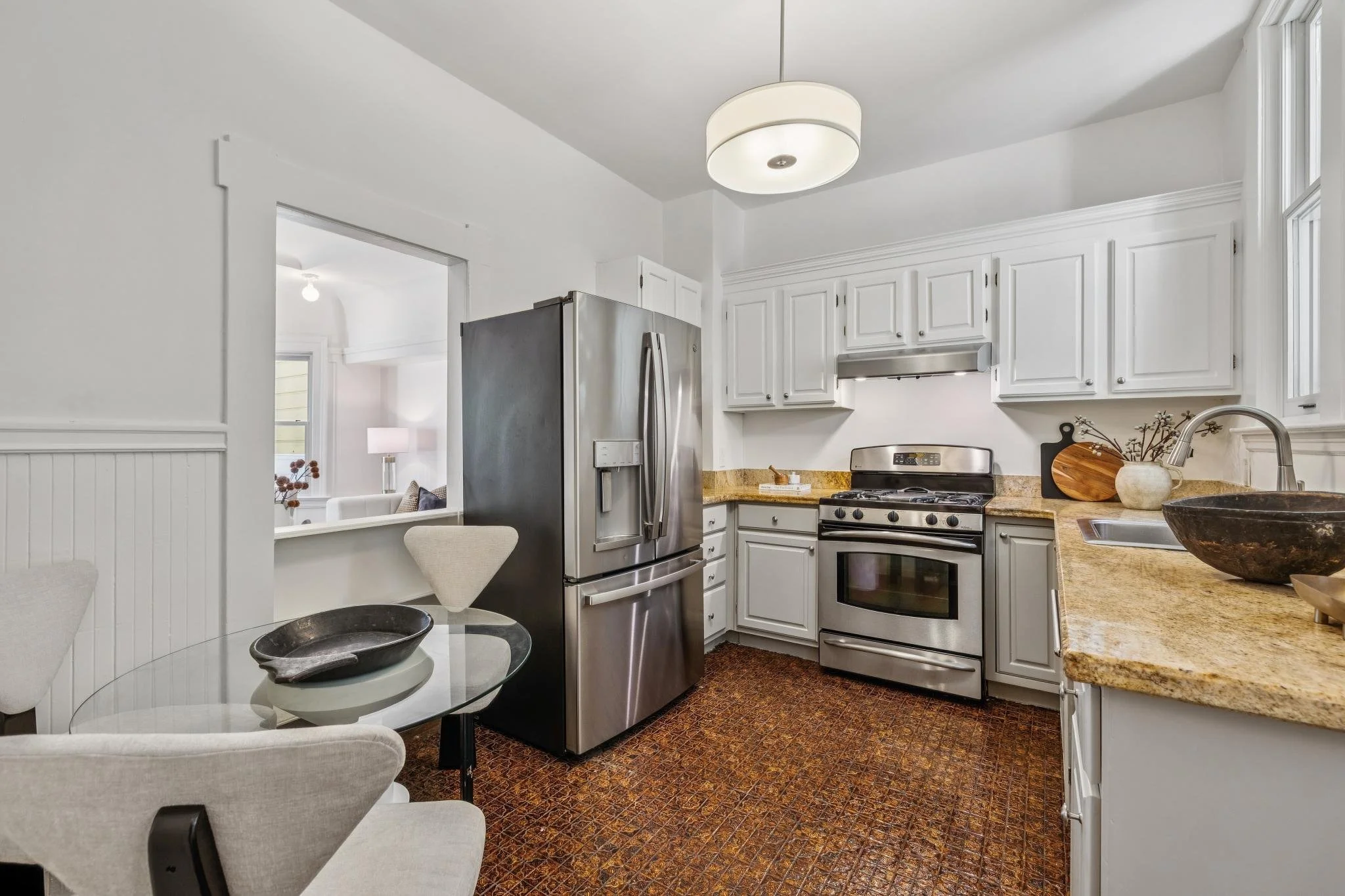View of a modern kitchen with white cabinets, stainless steel refrigerator, and stove. A round light fixture is on the ceiling, and there's a granite countertop with decorative items on it. A small dining table with white chairs is partially visible 