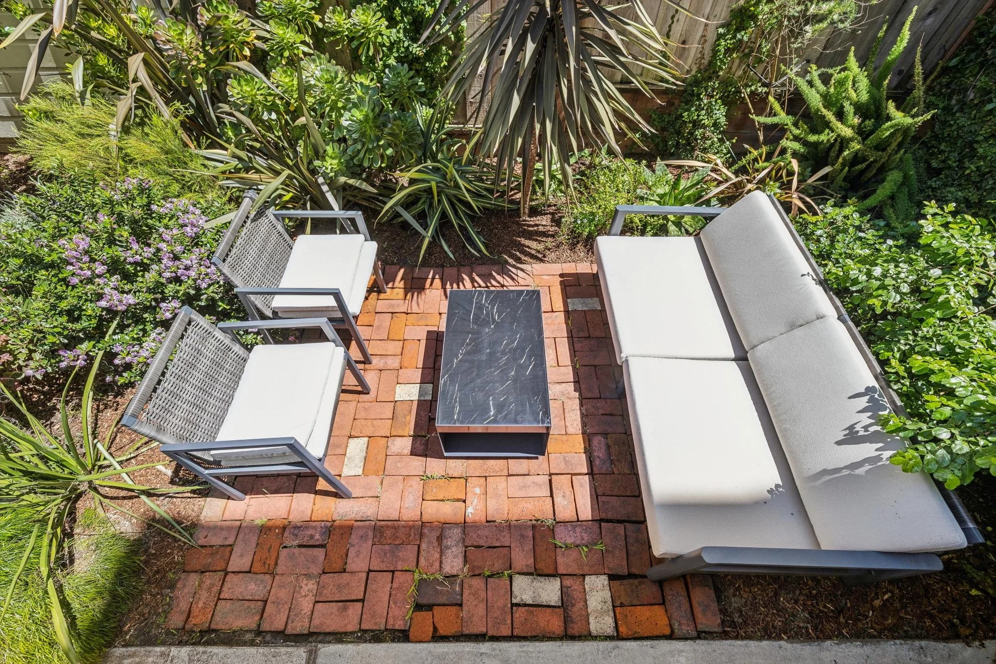 A small backyard patio with red brick paving, white outdoor furniture including two chairs and a bench, surrounded by green plants and bushes.