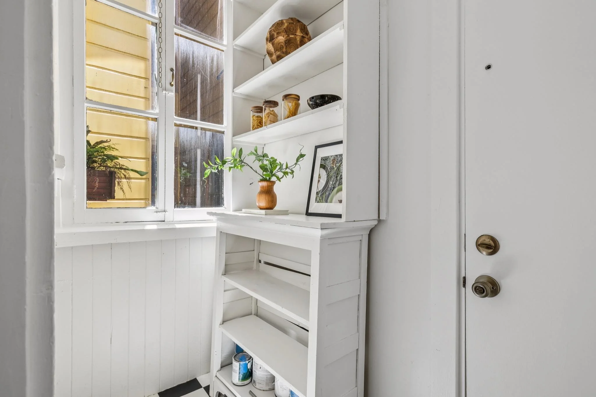 White wooden shelving unit with several shelves filled with jars, paint cans, and framed picture, located in a room with a window and a white door