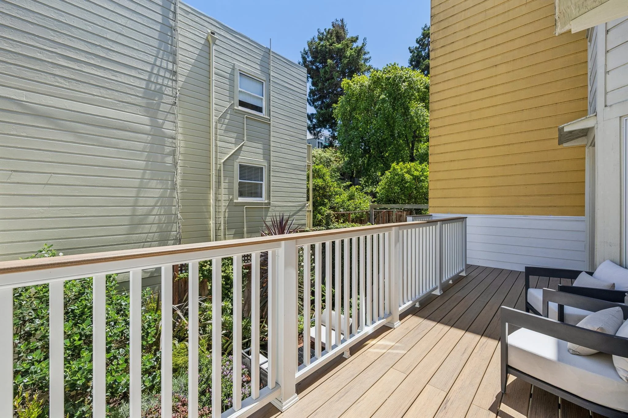 A view of a residential balcony with white railings, wooden flooring, and outdoor furniture including chairs. The balcony overlooks a garden with green bushes and trees, and neighboring houses are visible in the background under a clear blue sky.