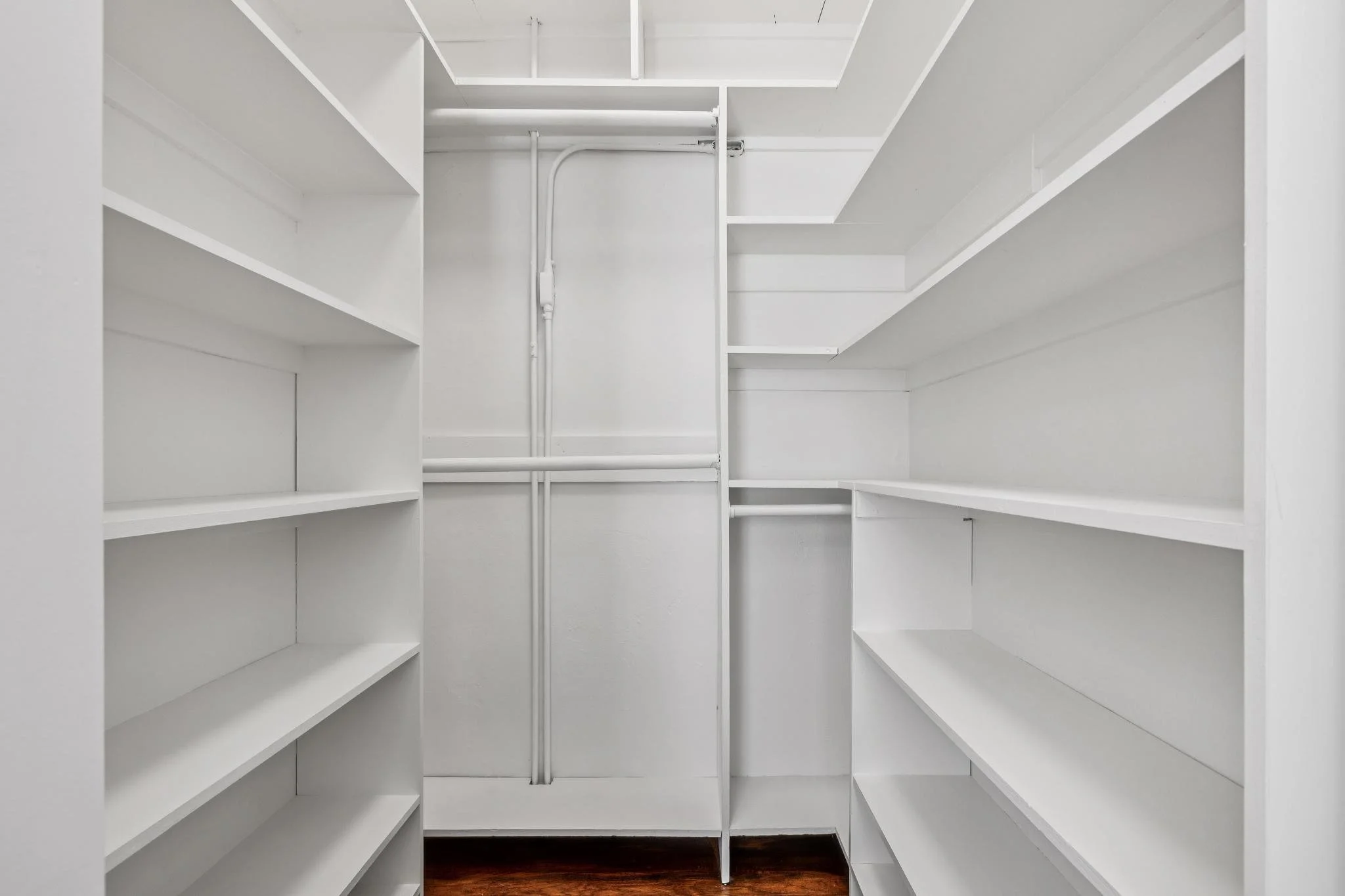 Empty white walk-in closet with shelves and hanging rods on both sides, floor made of dark wood.