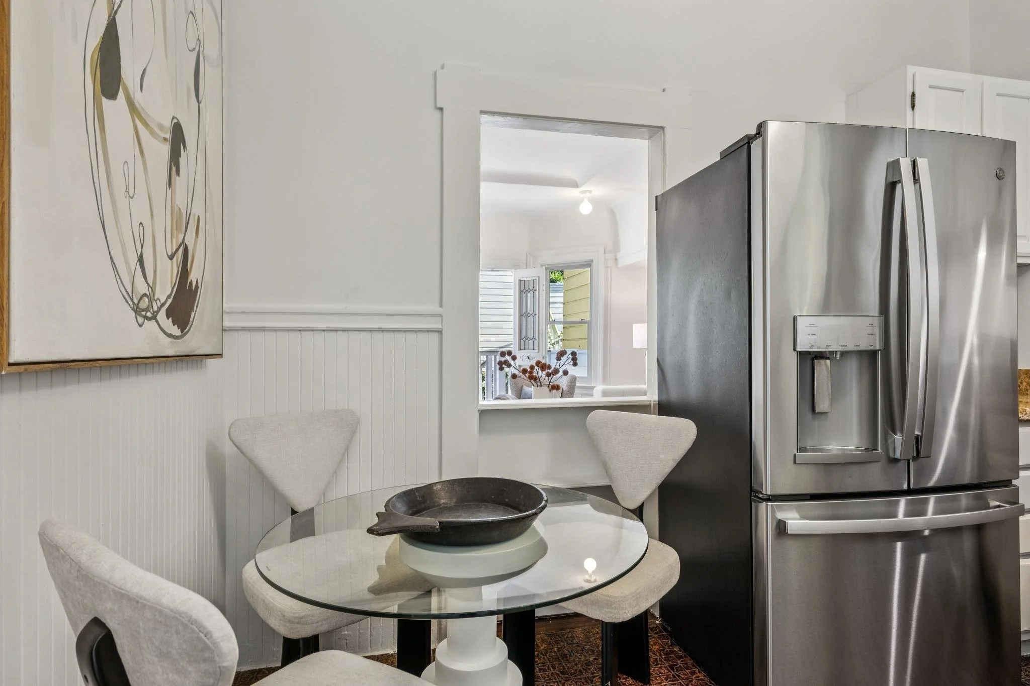 A kitchen view with a round glass dining table surrounded by four beige upholstered chairs, a black bowl on the table, a framed abstract art piece on the wall, a window with shutters showing a porch outside, and a stainless steel refrigerator.