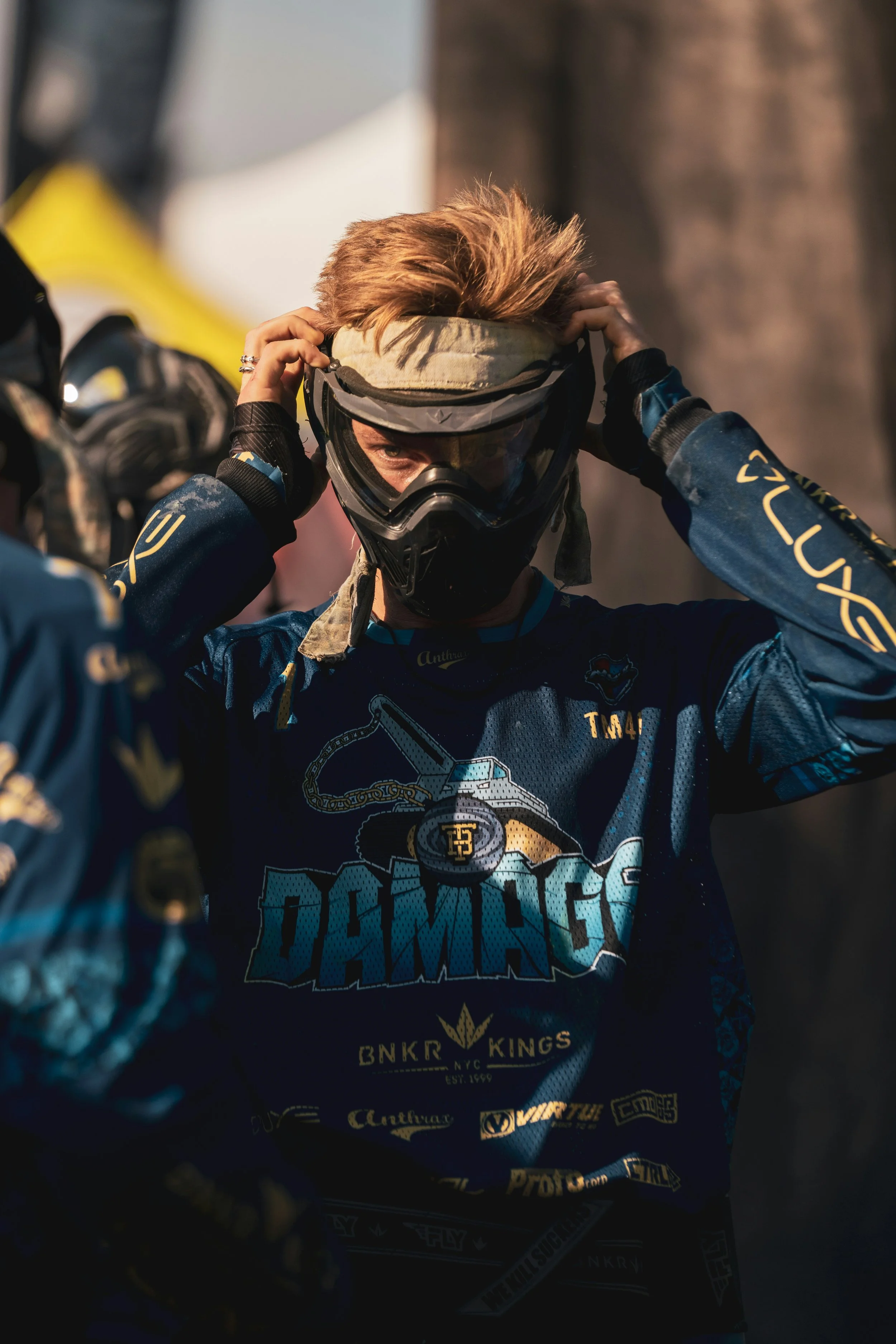 A paintball player adjusts their helmet, wearing a blue and black racing jersey. The player has blond hair and is surrounded by other team members or spectators, with sunlight highlighting their face and helmet in central alberta.