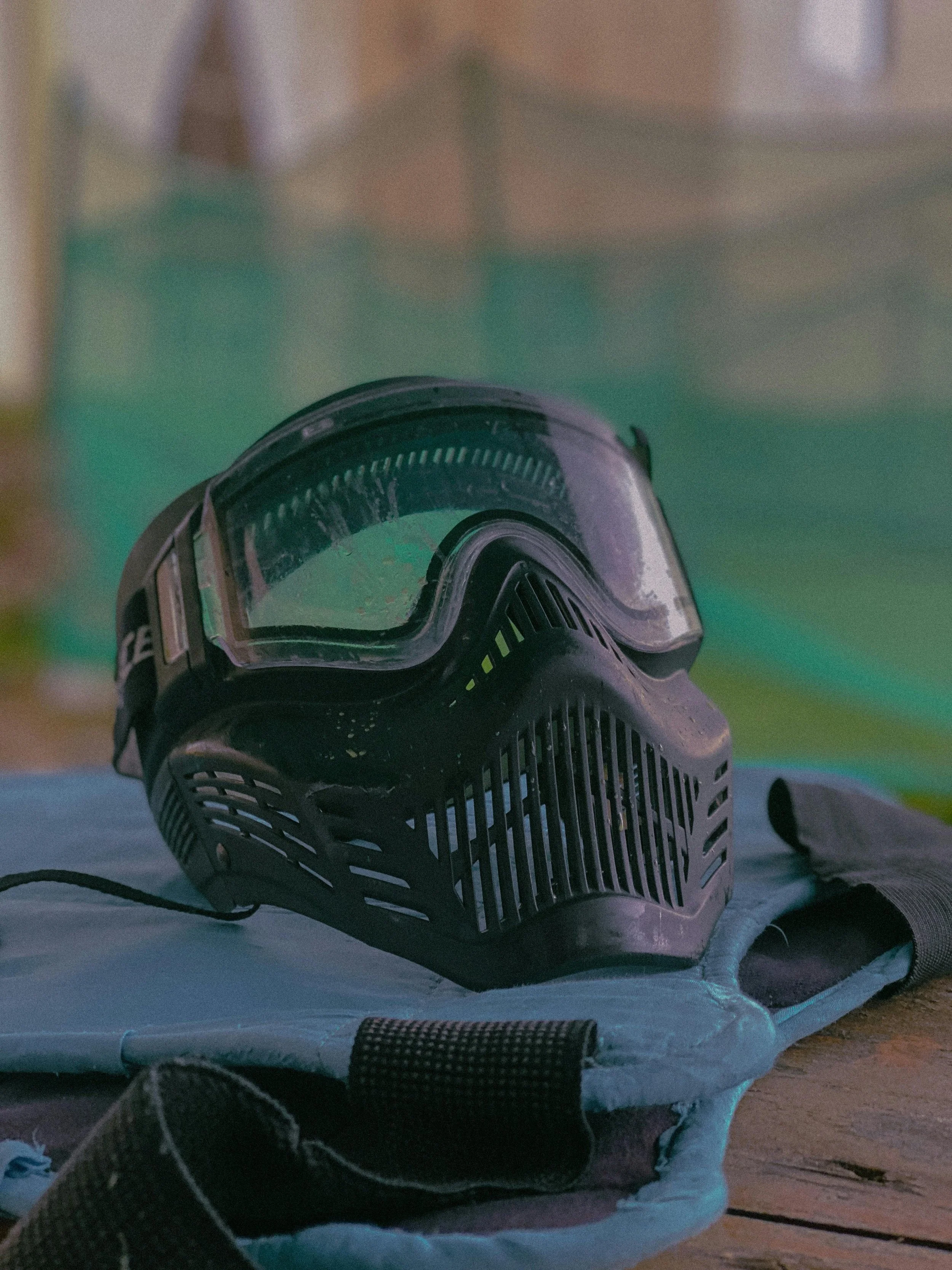 A pair of black ski goggles with a clear lens and yellow accents resting on a gray padded surface next to a black strap with a Velcro closure. The background is blurred with soft, warm lighting in central alberta.