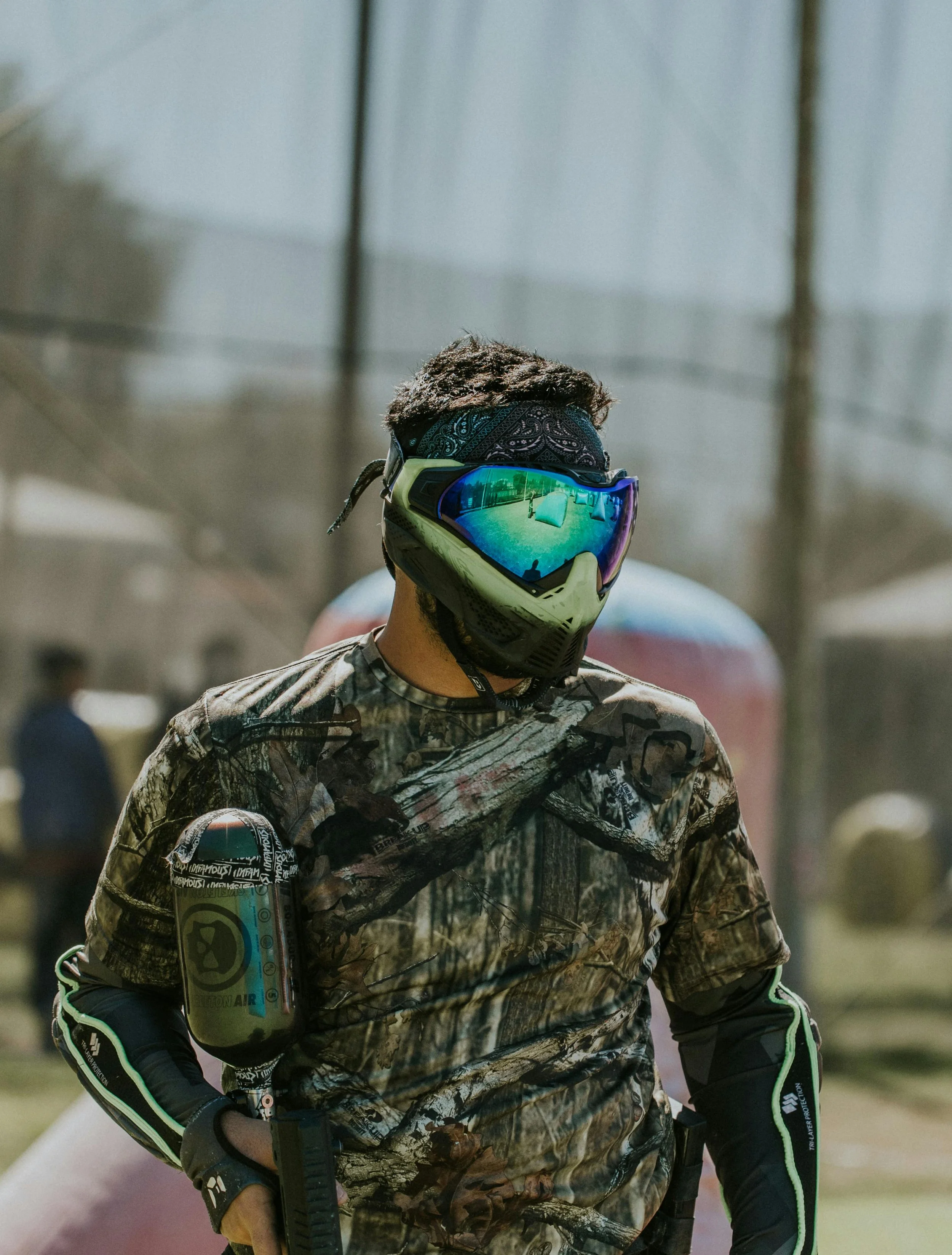 A person wearing camouflage clothing, a bandana, and reflective goggles, holding a paintball gun with a camouflage design in an outdoor setting.