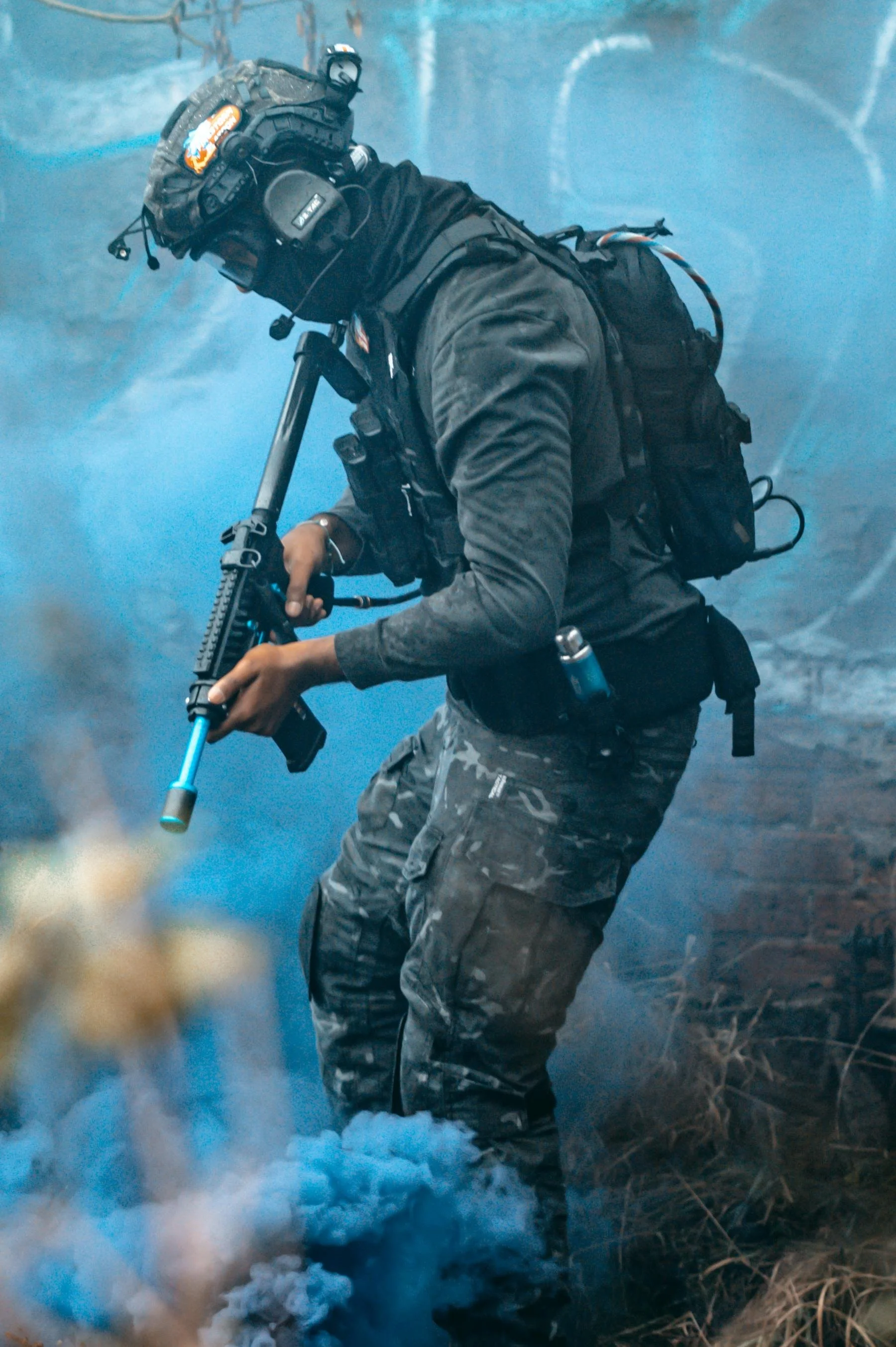 A person dressed in tactical gear and camouflage pants, wearing a helmet with a headset, is holding an airsoft gun, surrounded by blue smoke in an outdoor environment in central alberta.