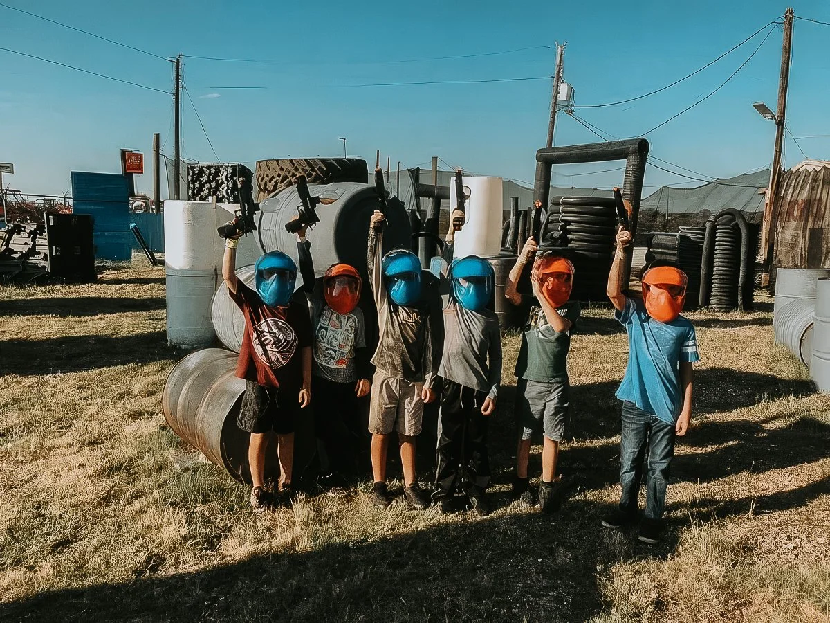 Six children wearing protective masks and holding toy guns in a paintball or laser tag setting, with large protective tubing, barrels, and industrial structures in the background in central alberta.