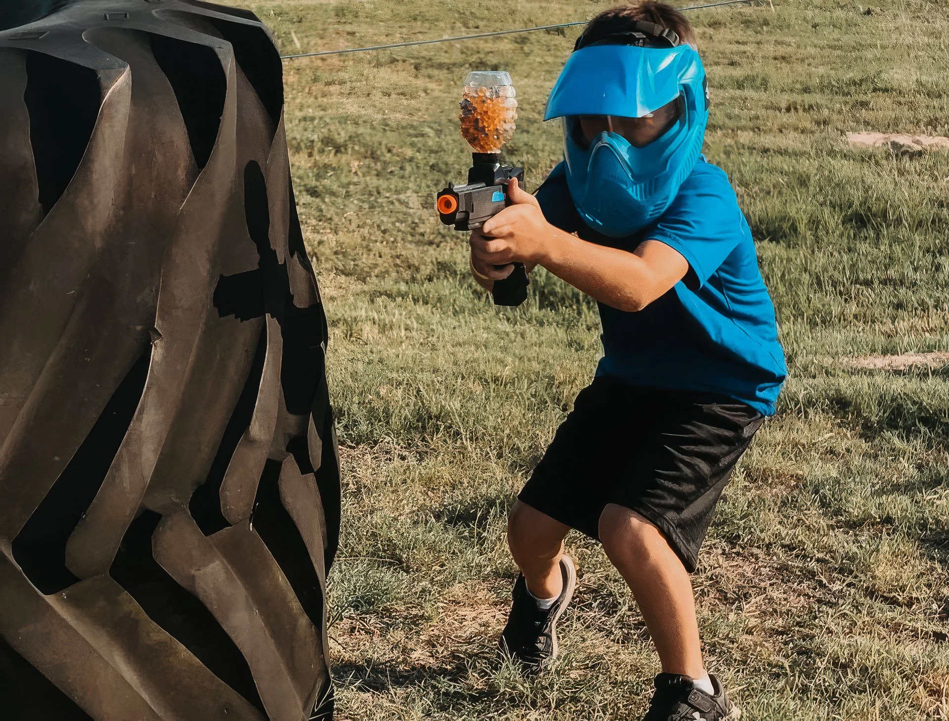 Child in blue shirt and shorts wearing a blue protective mask and helmet, holding a gellyball blaster, crouched behind a large black tire in an outdoor grassy area in central alberta.