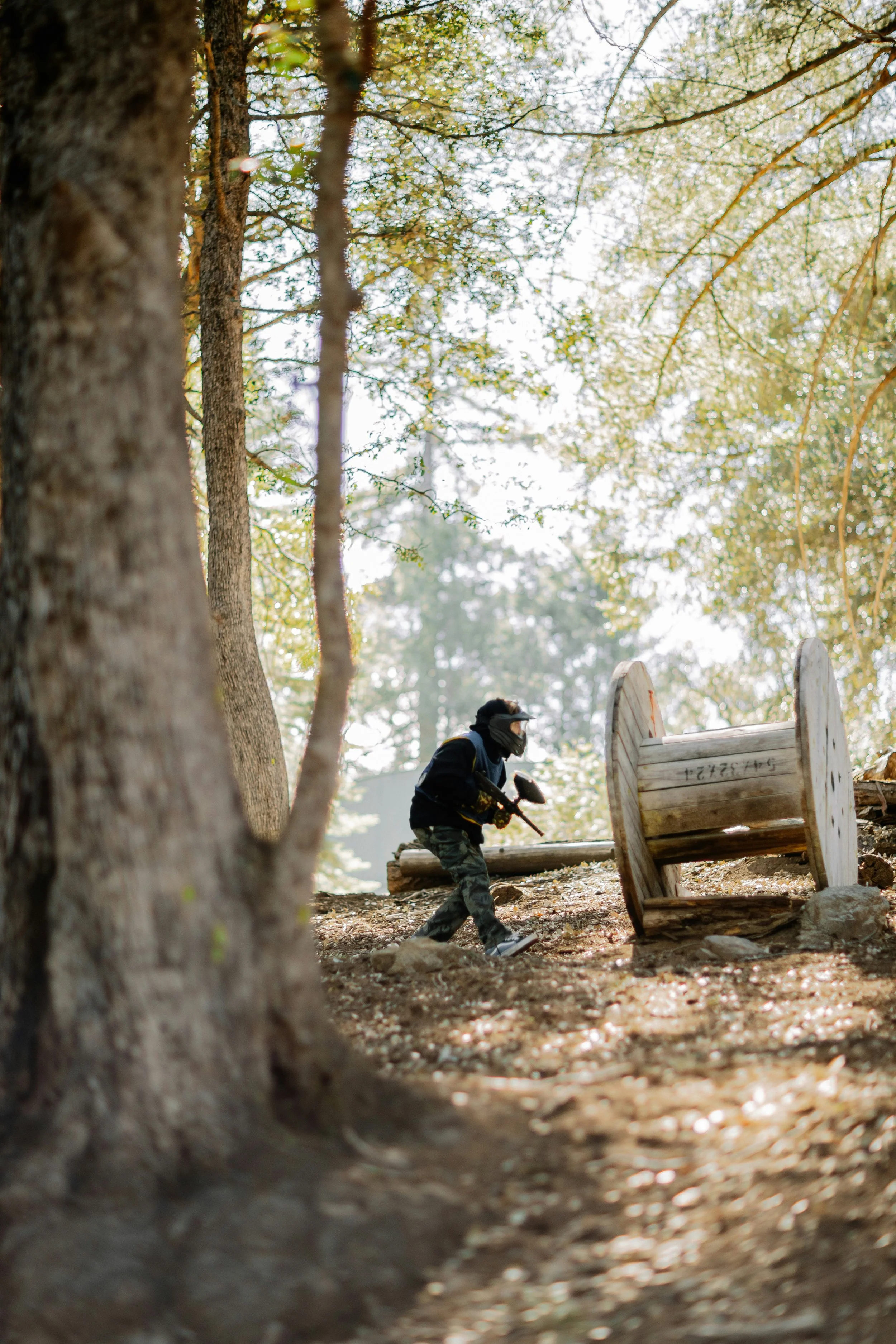 A person wearing camouflage pants, a black jacket, a mask, and gloves is holding a paintball gun while playing paintball by a wooden spool replica in a forest with tall trees and sunlight filtering through the leaves in central alberta.