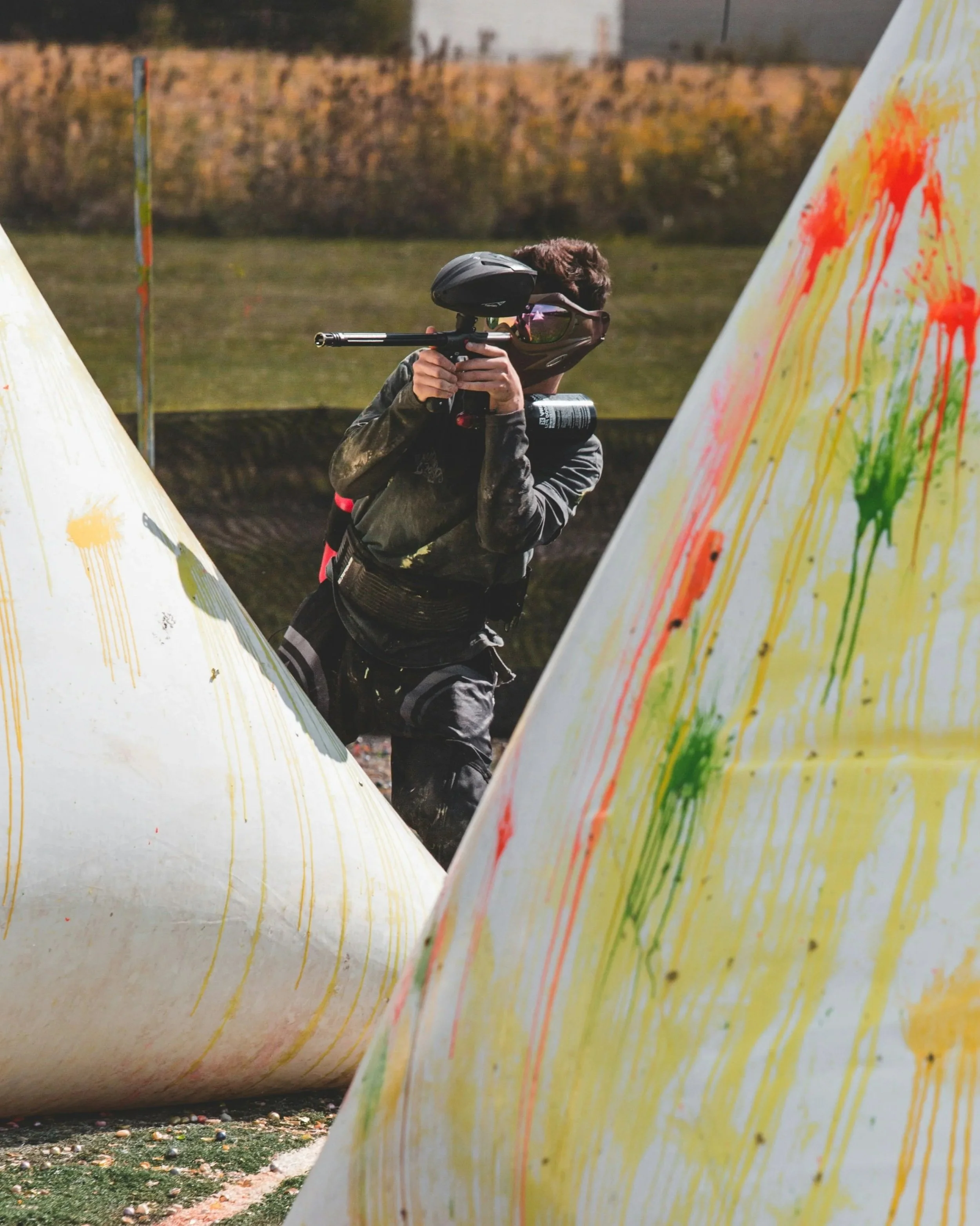 A person wearing a mask, sunglasses, and camouflage shirt aiming a paintball gun behind a painted obstacle at an outdoor paintball field in central alberta.