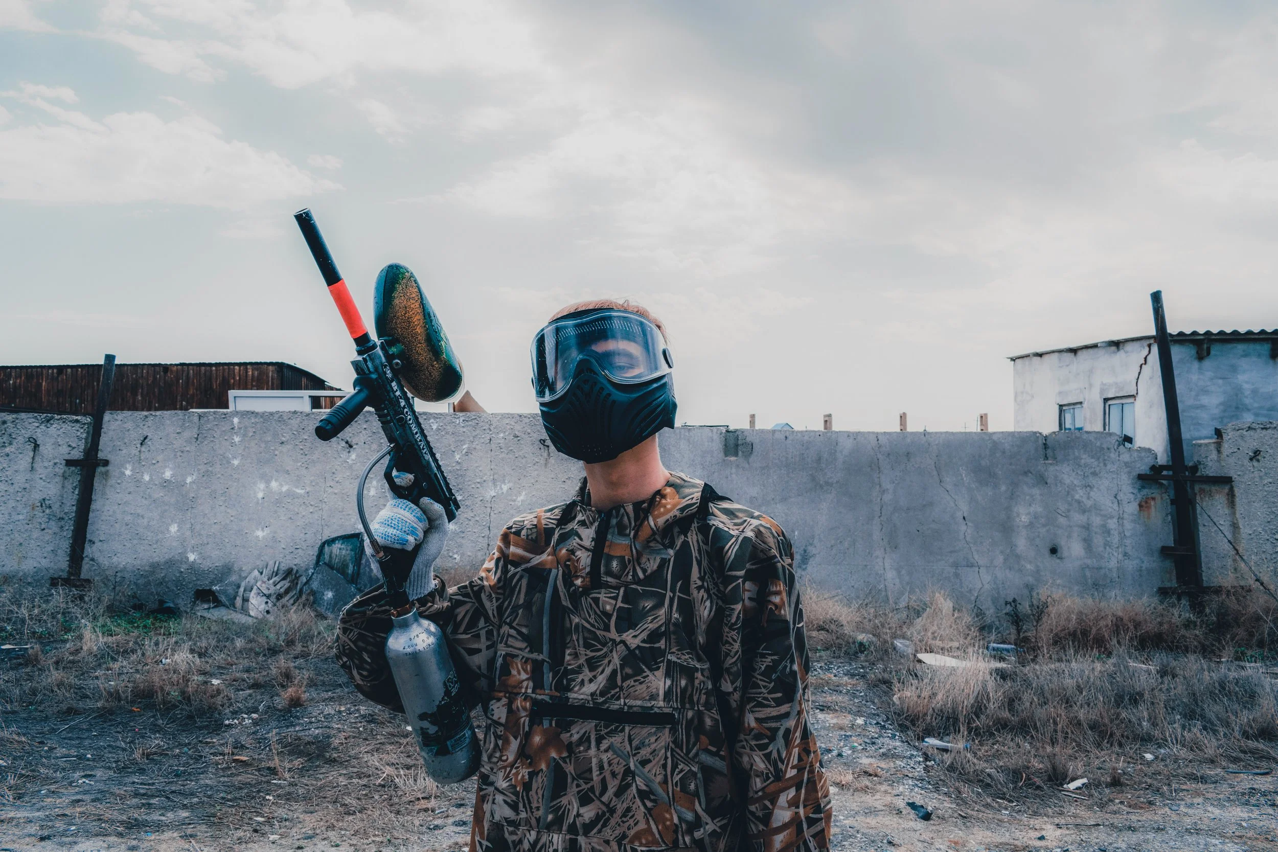 Person wearing a paintball mask and camouflage jacket holding a paintball gun outdoors near a concrete fence in central alberta