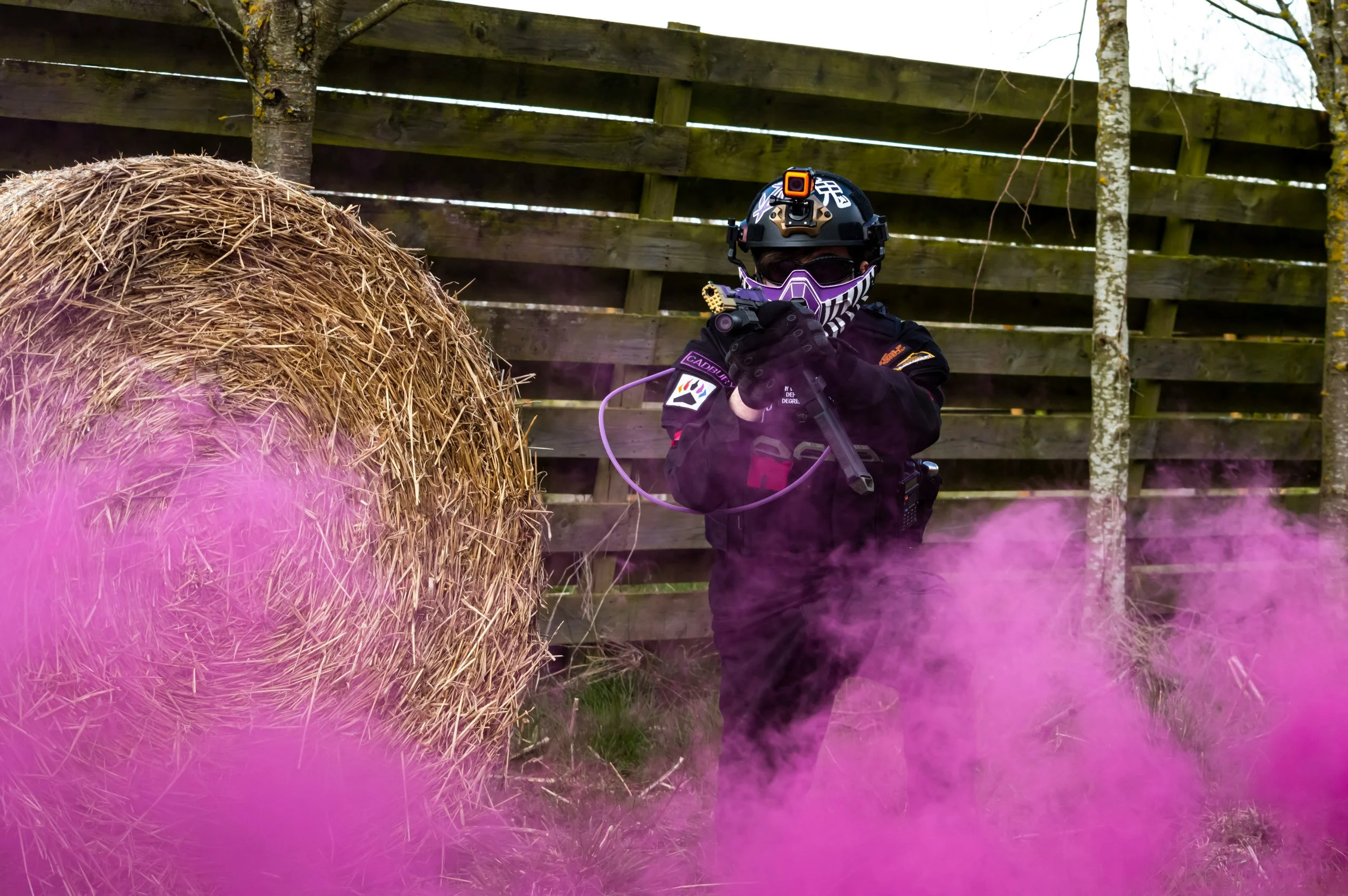 A person wearing a helmet with a headlamp, sunglasses, and a striped face covering, holding a device near a large round hay bale, with pink smoke and a wooden fence in the background in central alberta