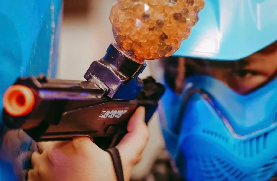 A person wearing protective gear, including a blue face mask, face shield, and gloves, aiming a toy gun with a large, textured, orange and black foam dart at a blue wall in central alberta