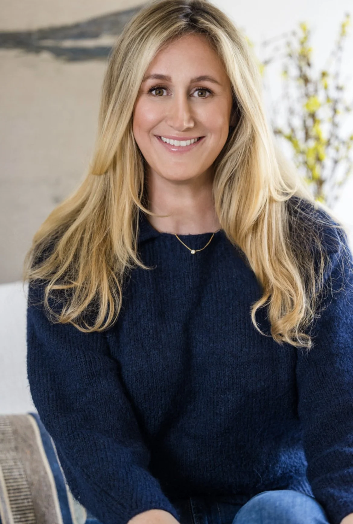 A woman with long blonde hair smiling, wearing a navy blue sweater, sitting indoors with a plant in the background.