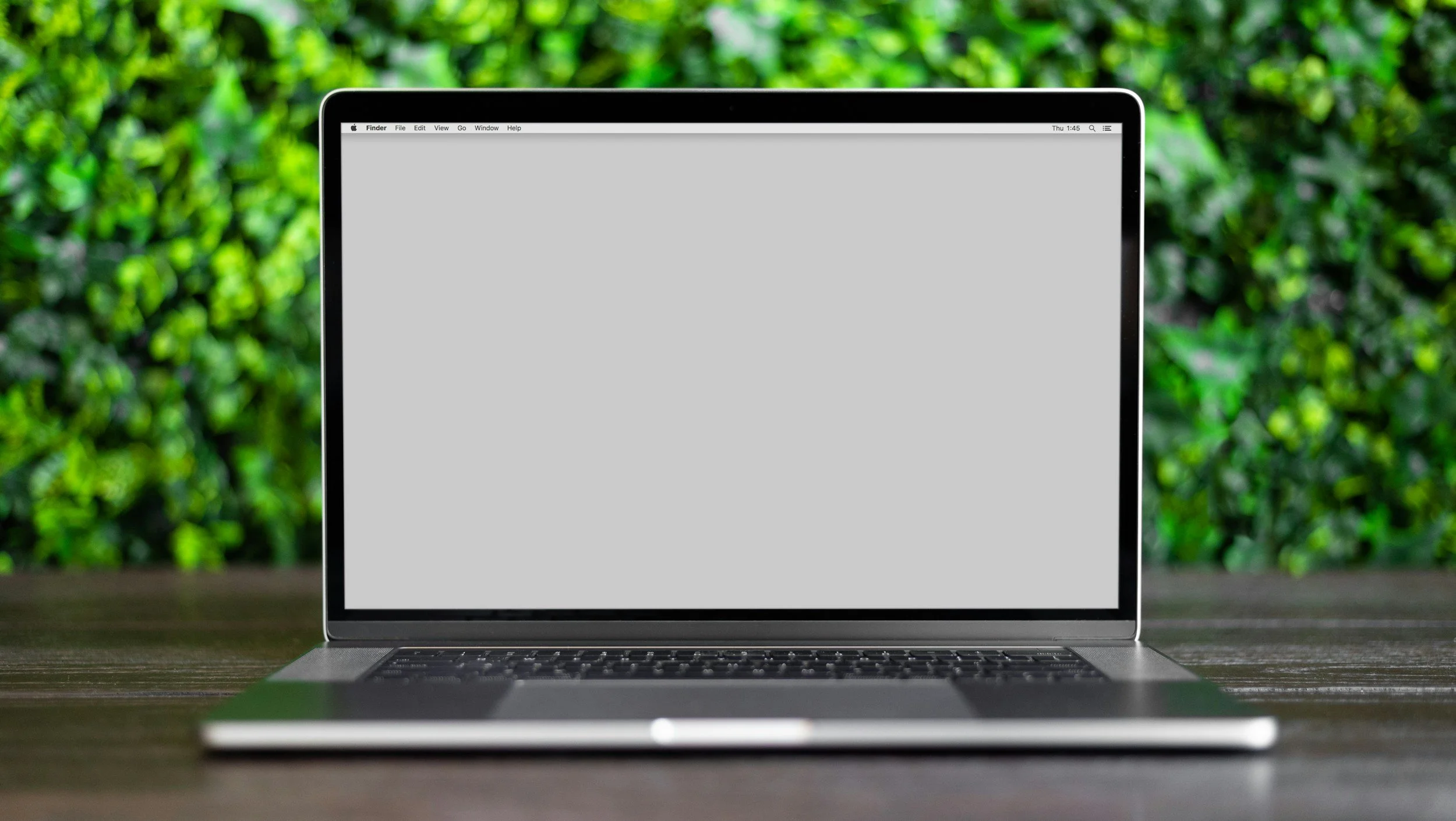 Laptop with a blank gray screen on a wooden table outdoors with green bushes in the background, virtual therapy.