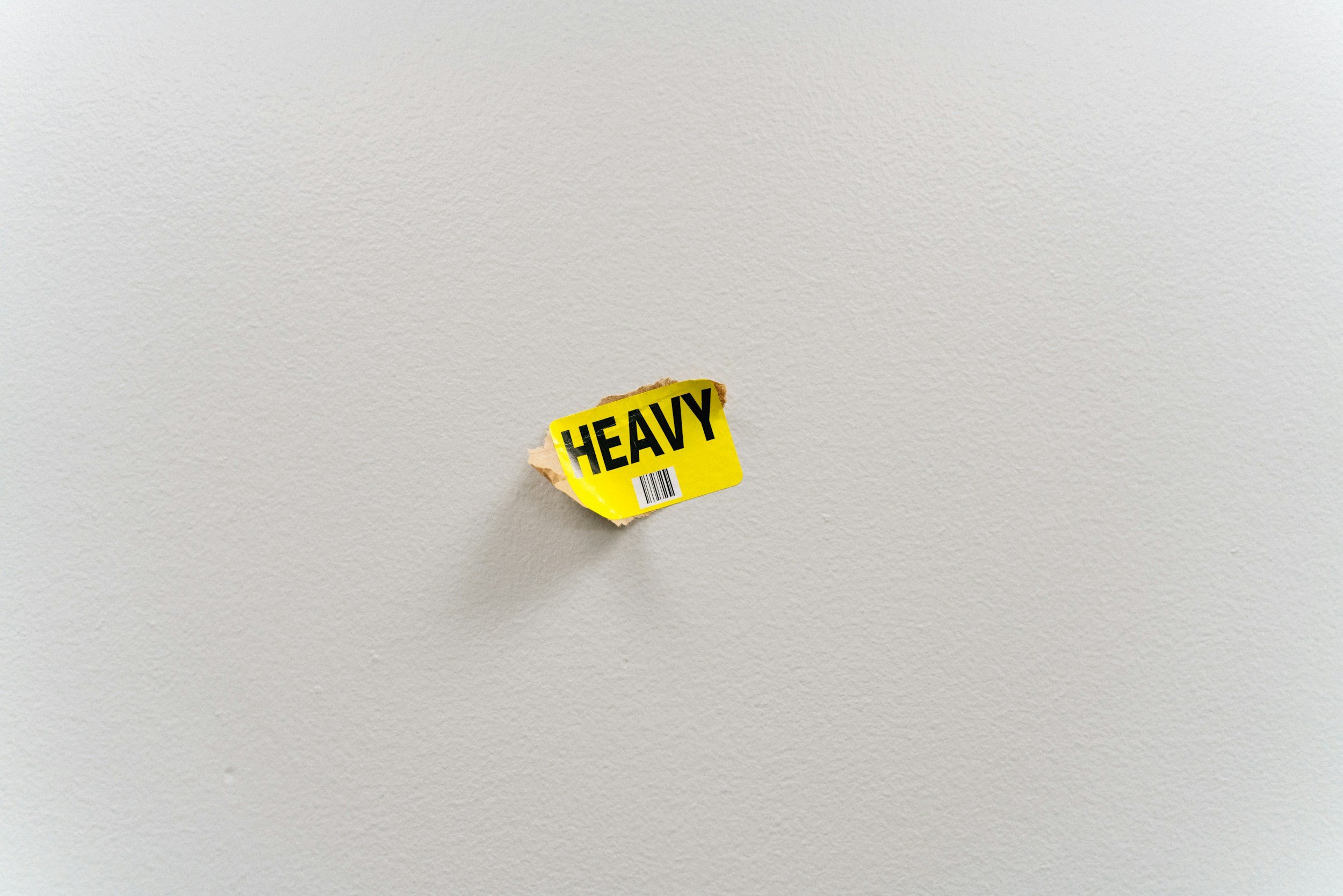 Yellow sticker with the word 'HEAVY' attached to a small torn piece of paper on a plain white wall.