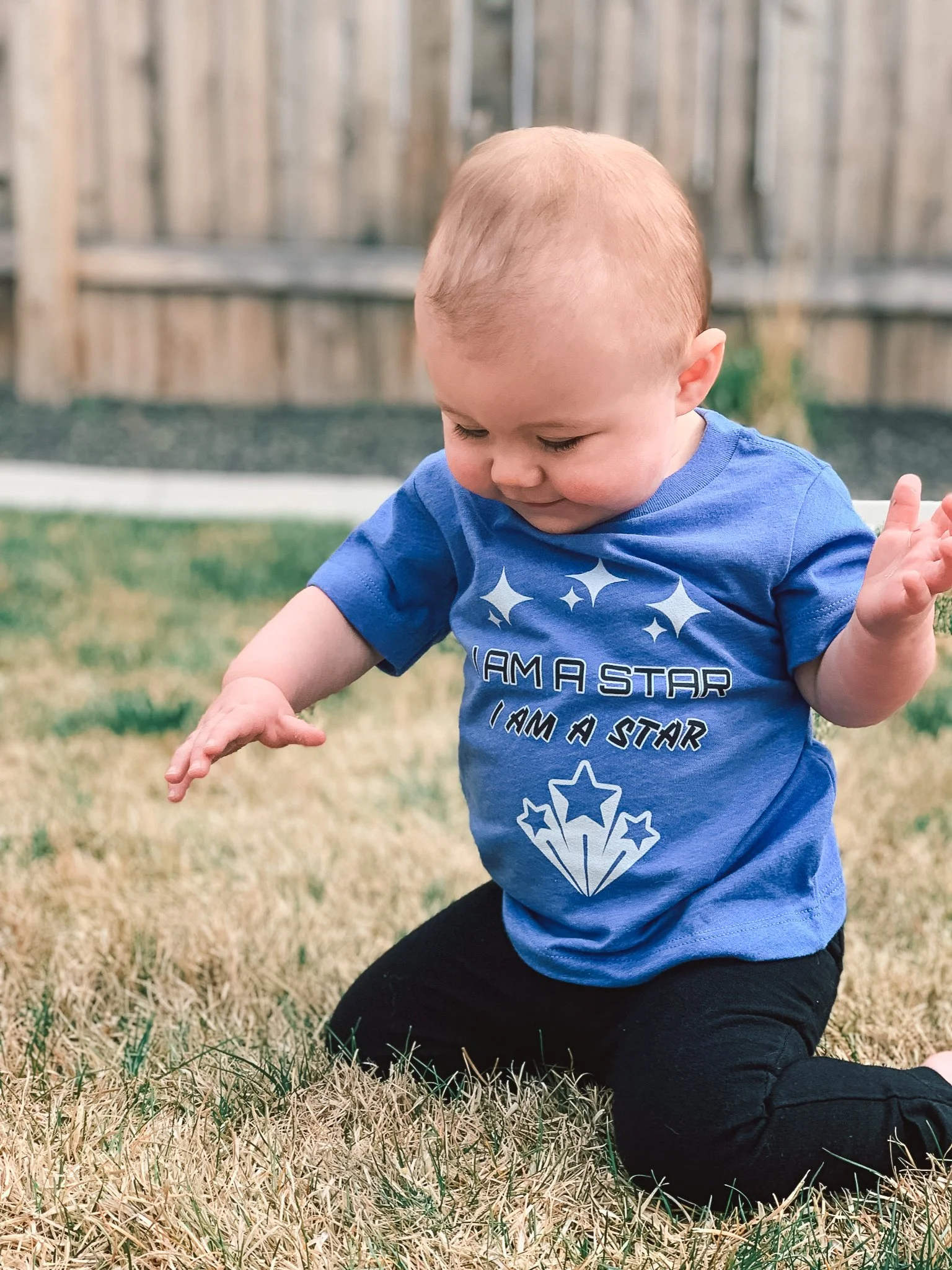 A young child with light hair wearing a blue T-shirt with star graphics and black pants, kneeling on dry grass in a backyard with a wooden fence in the background.