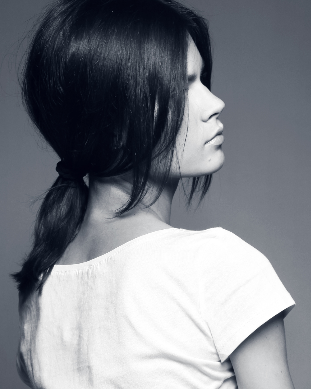 Black-and-white profile photo of a woman with dark hair tied in a low ponytail, wearing a white t-shirt, against a plain gray background.