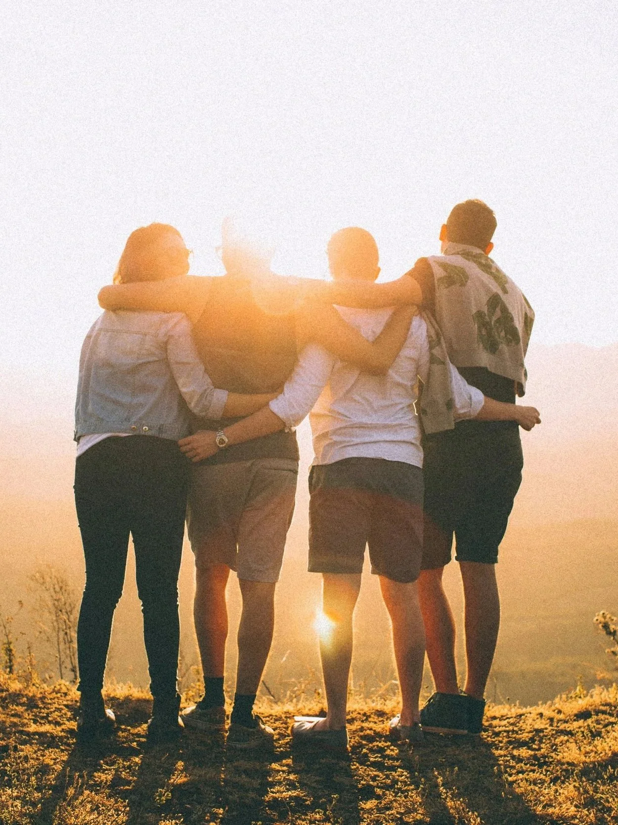 Four friends walking together at sunset with arms around each other, enjoying friendship and field scenery.