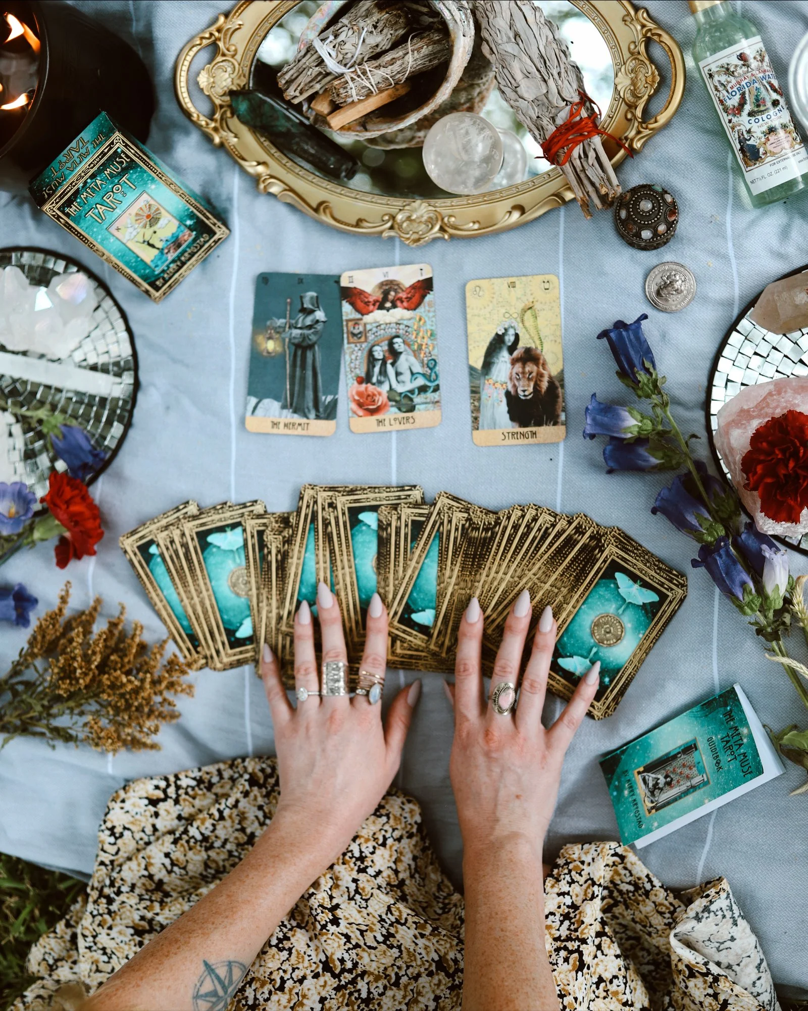 A person with tattoos and rings on their fingers is shuffling Tarot cards on a table covered with a light blue tablecloth. The cards display mystical imagery with titles like "The Hermit," "The Lovers," and "Strength." Surrounding the cards are flowe