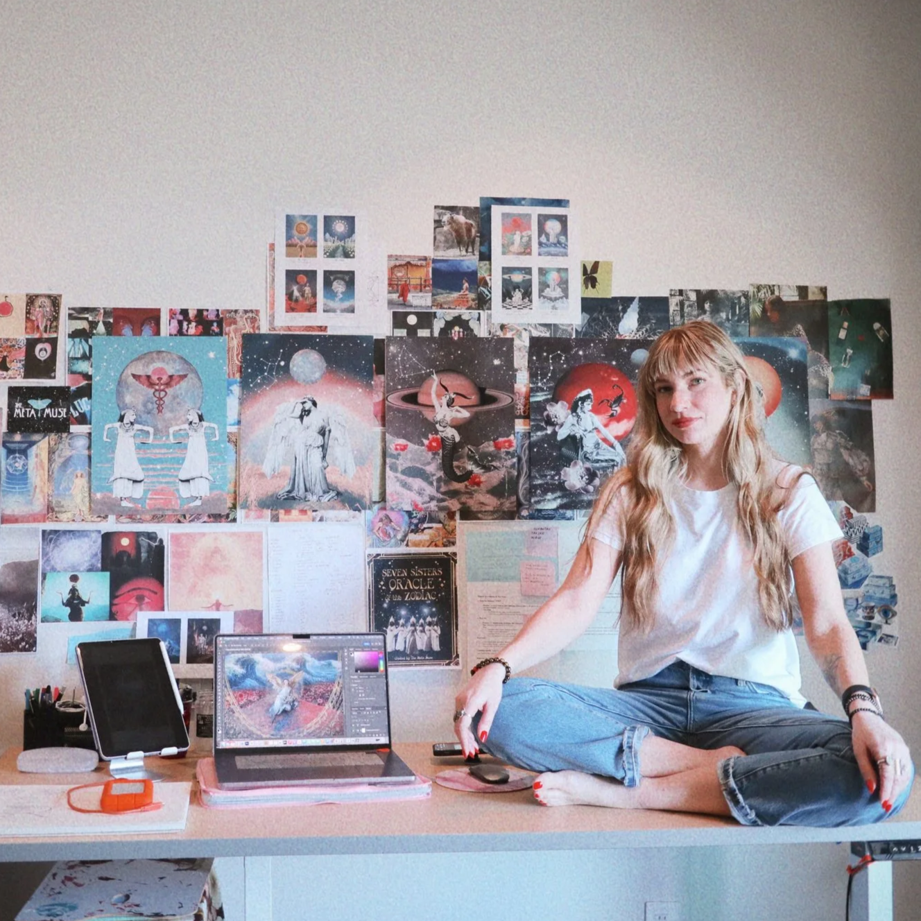 A young woman with long blond hair sitting on a desk in front of a wall decorated with colorful artwork and posters, with a laptop and tablet on the desk.
