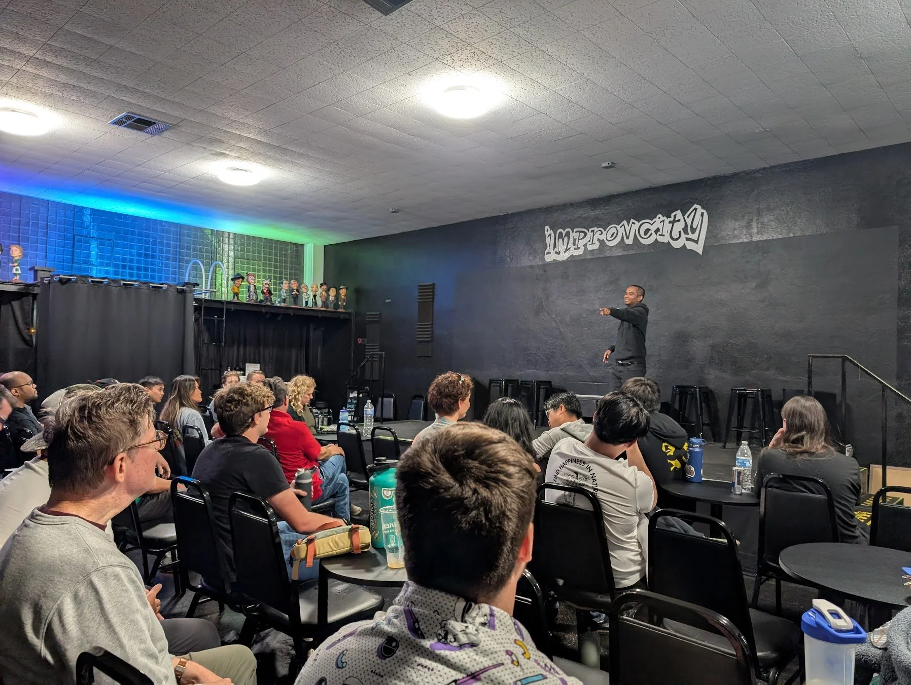 A man standing on stage speaking to an audience in a conference room with black walls and ceiling, decorated with cutouts spelling 'improvcity,' with several puppets or figurines on a shelf along the back wall, and audience members sitting and listening.