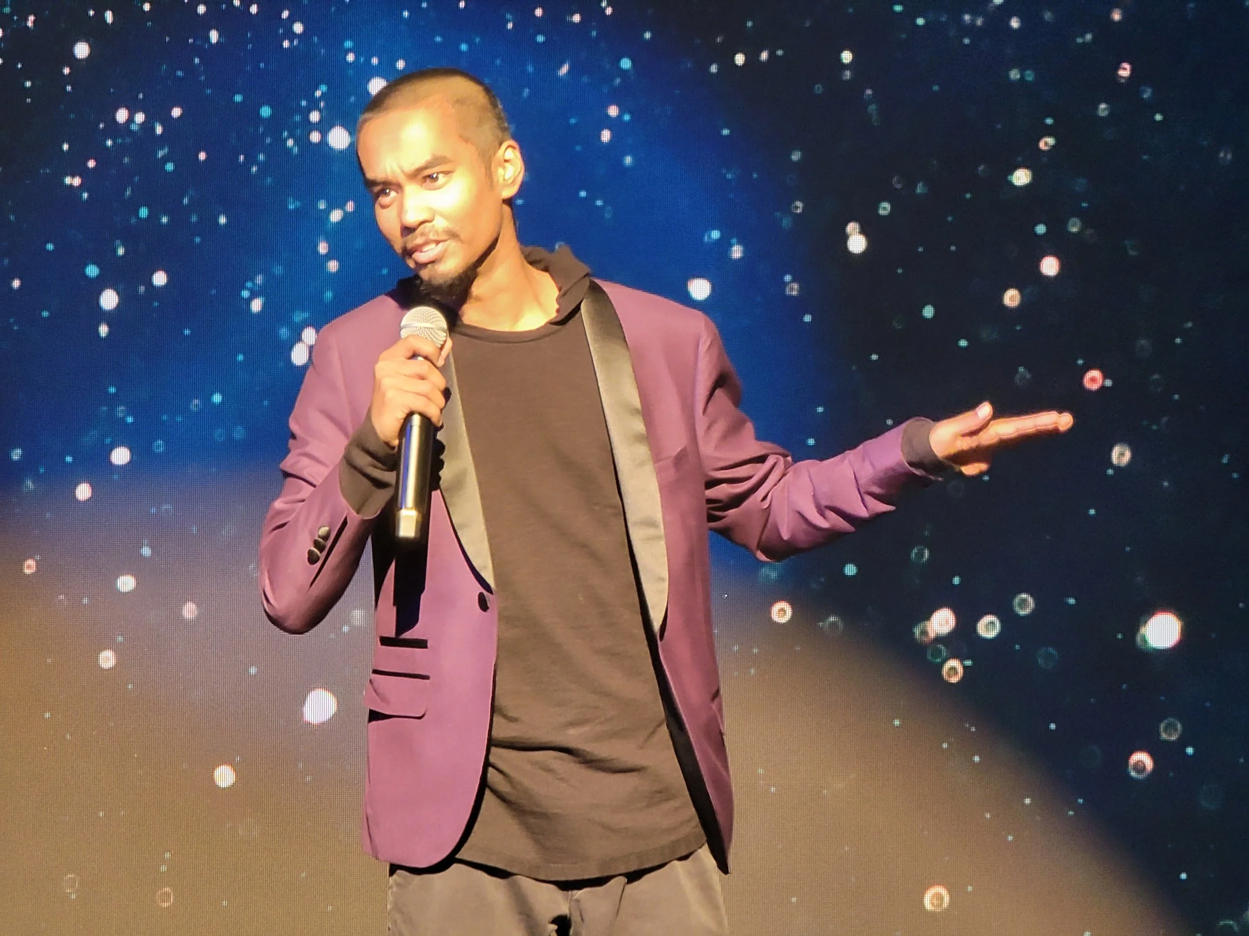A man wearing a purple jacket and brown shirt holding a microphone on stage with a background of blue and white light dots.