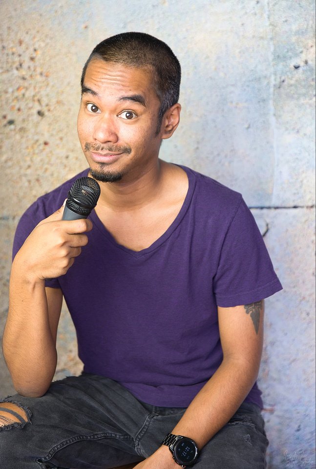 A man with short hair and a goatee, wearing a purple T-shirt and ripped jeans, sitting against a textured wall, holding a microphone and looking at the camera.