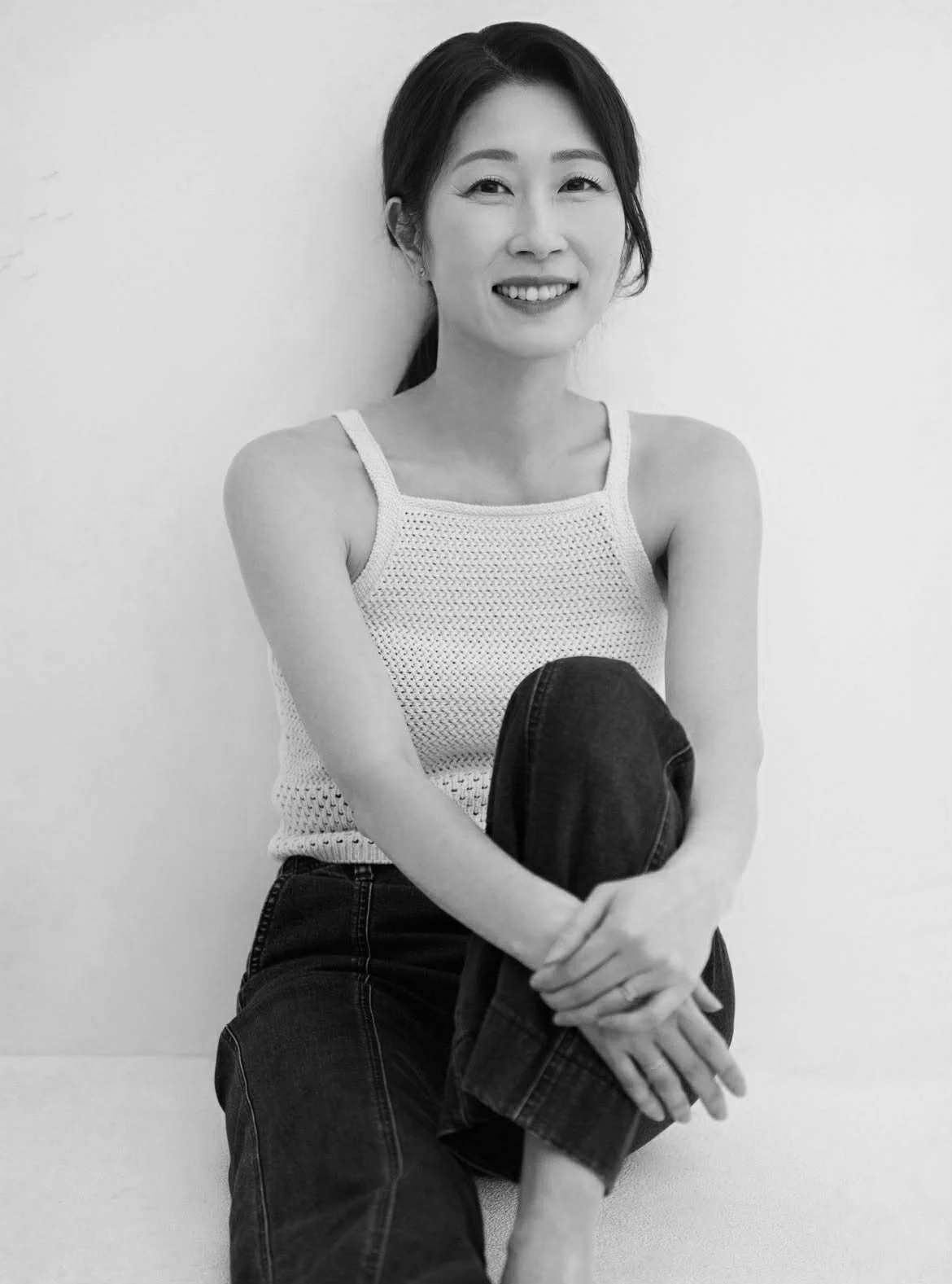 Black and white photograph of a smiling Asian woman with dark hair tied back, wearing a sleeveless knit top and dark jeans, seated with one knee bent and resting on her lap, against a plain wall.