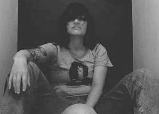 Person with dark hair and glasses sitting on the floor in a small enclosed space, wearing a t-shirt and jeans.