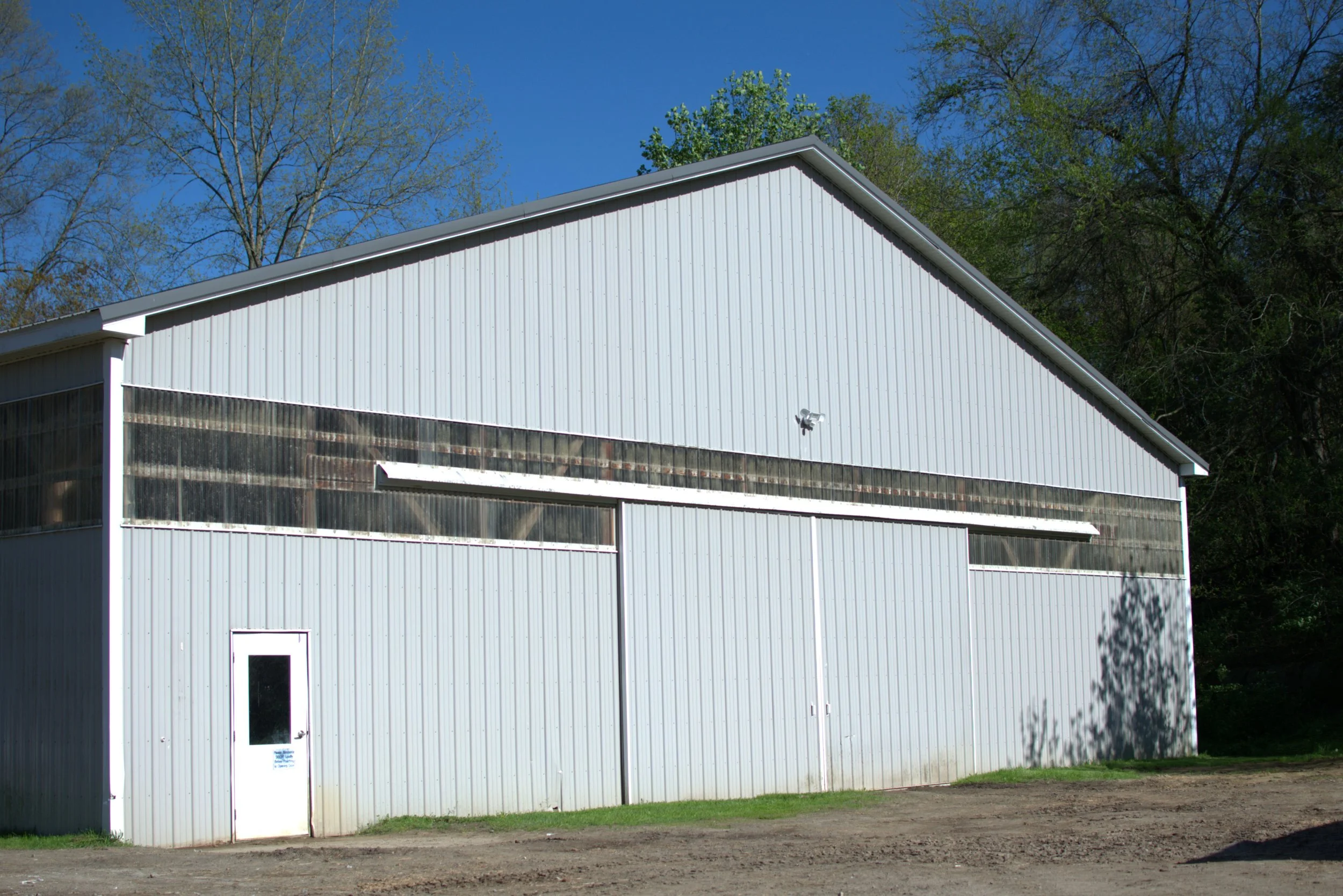 White metal building with a sloped roof, a glass window, and a white door, set against a backdrop of leafless and green trees and a clear blue sky.