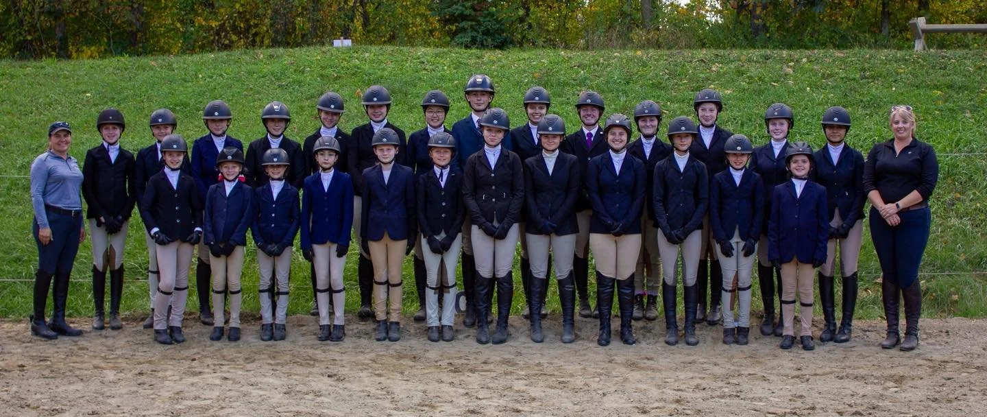Group of young equestrians in riding attire, including helmets and riding boots, posing outdoors with two adult women, on a sandy riding arena with greenery and trees in the background.